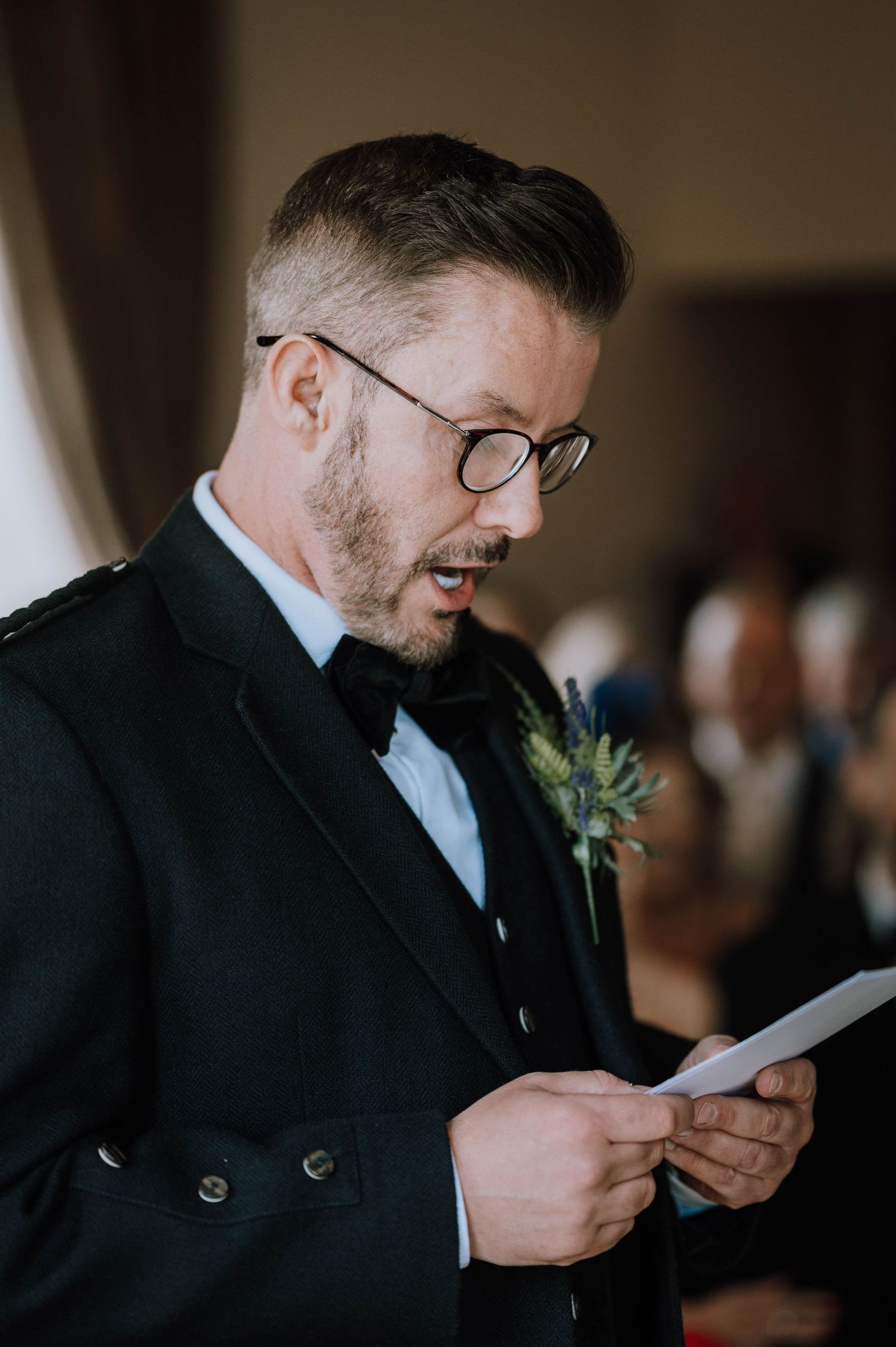 Logie House Wedding Photography. A person in a black suit and glasses reads a letter during an emotional moment at a wedding ceremony.