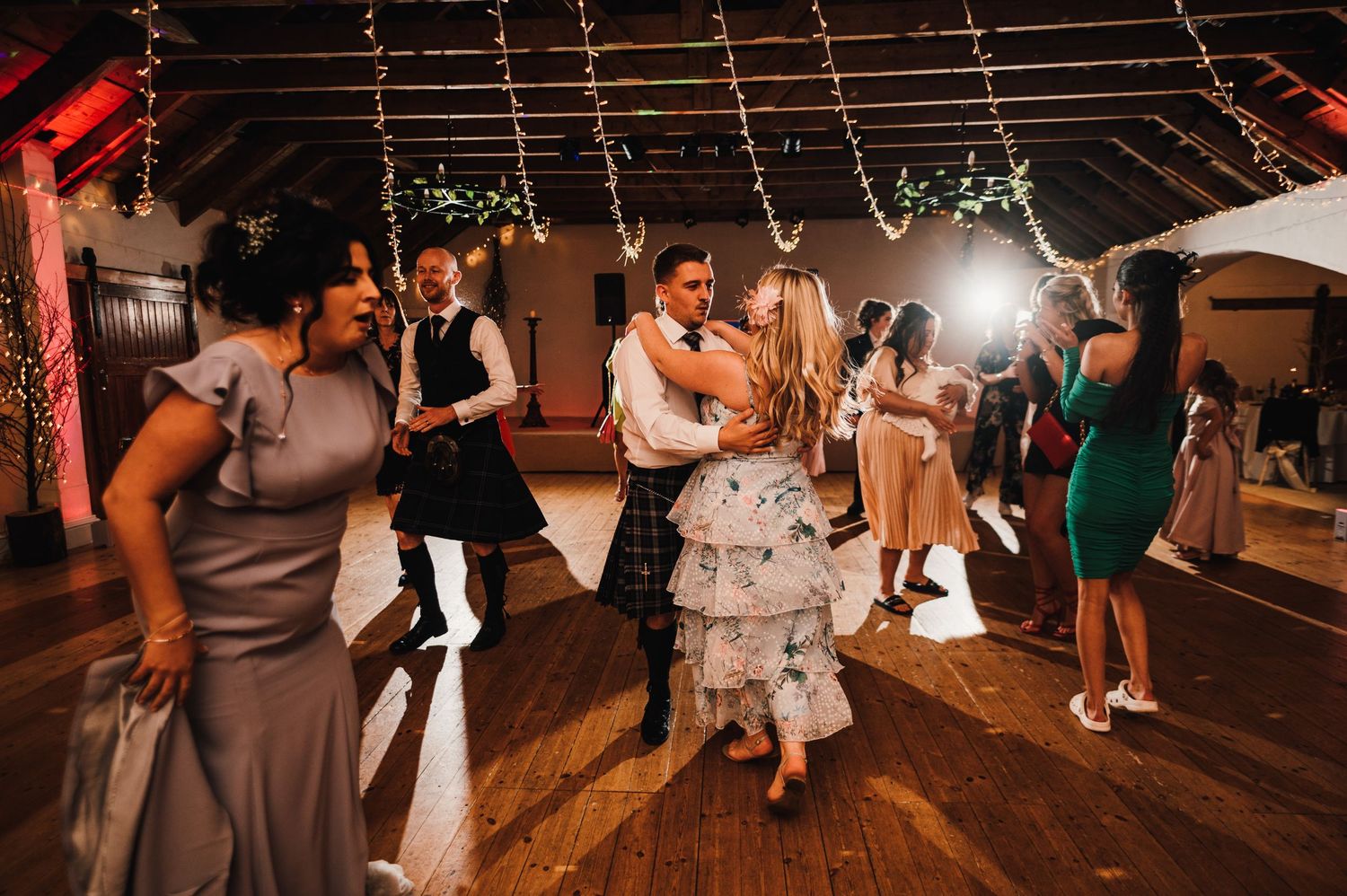 Couples dancing and celebrating on a rustic wooden dance floor under warm lighting at an evening reception.