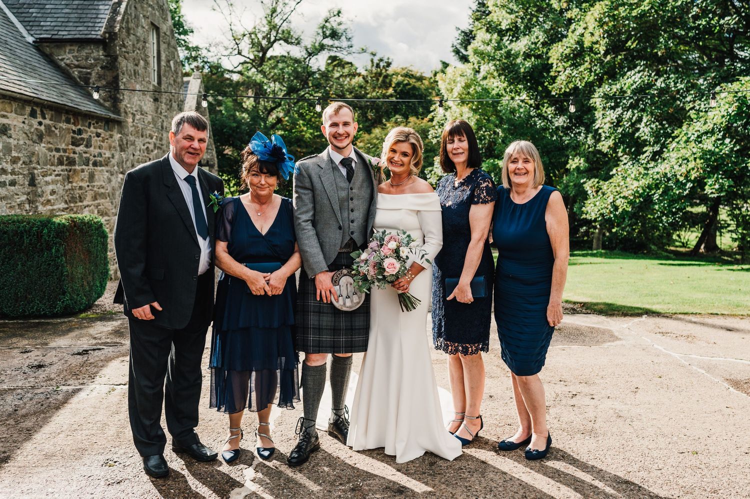 Wedding group photo with guests in formal attire standing outside stone church.