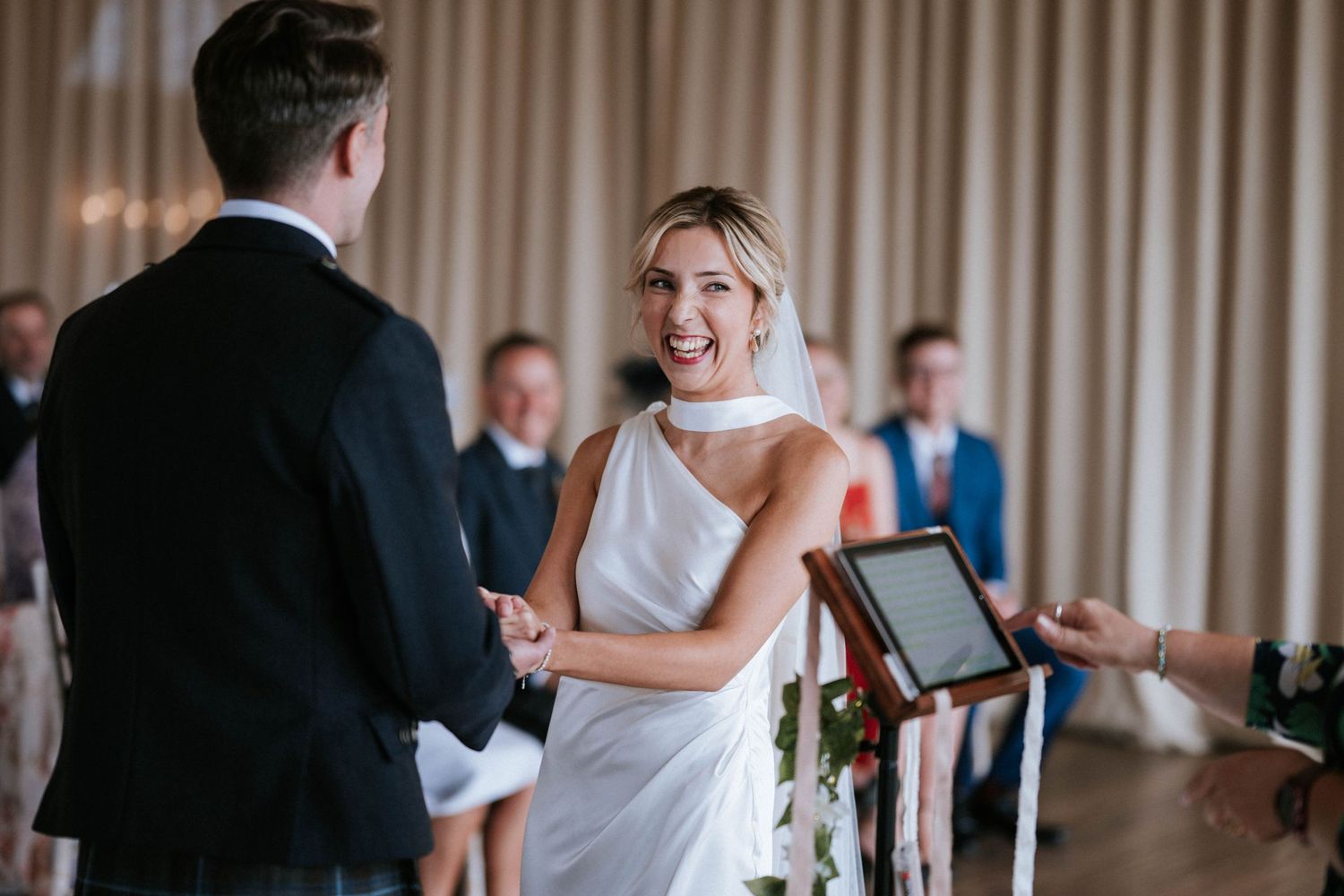 Wedding ceremony in progress inside venue with beige curtains and natural lighting.