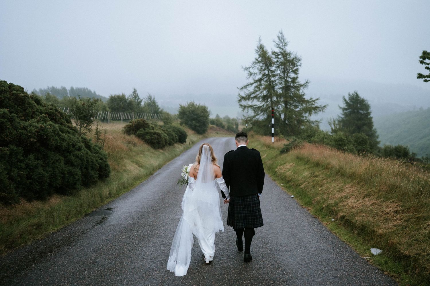 A couple walks along a misty country road in Scotland wearing wedding attire on their special day.