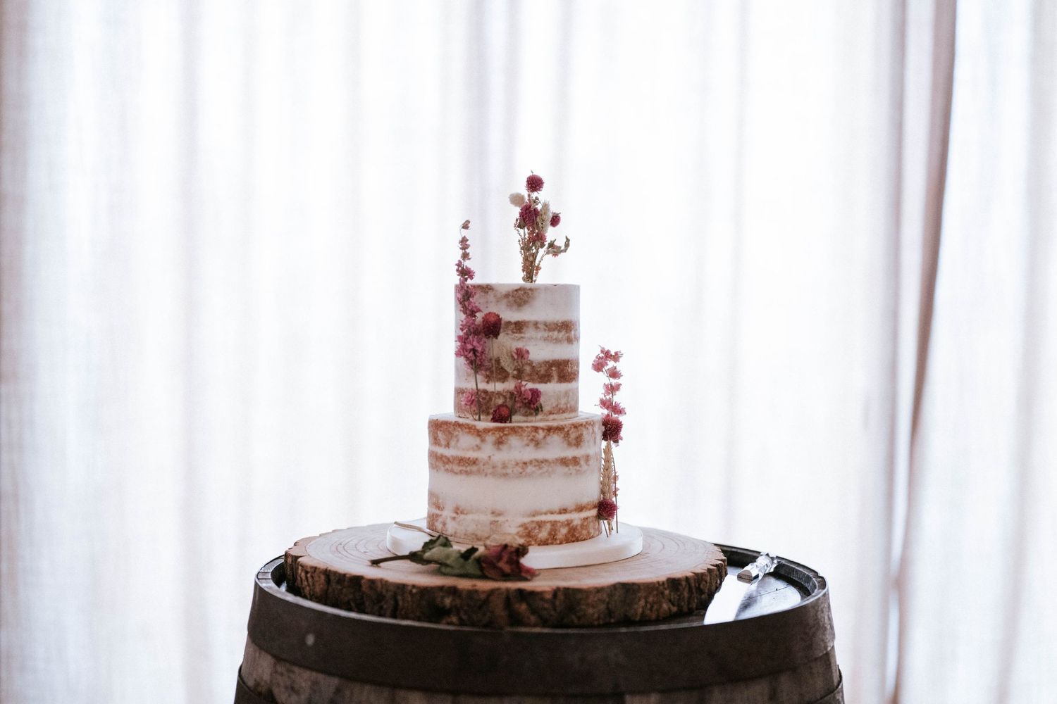 Semi-naked wedding cake with red berries displayed on rustic wooden barrel.