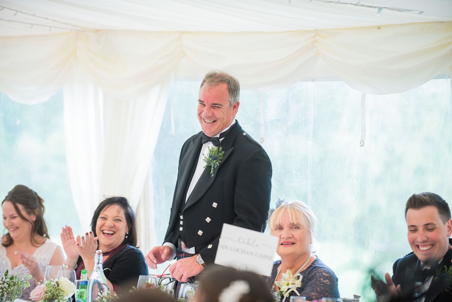 Wedding reception guests seated at tables under a white tent during celebrations.