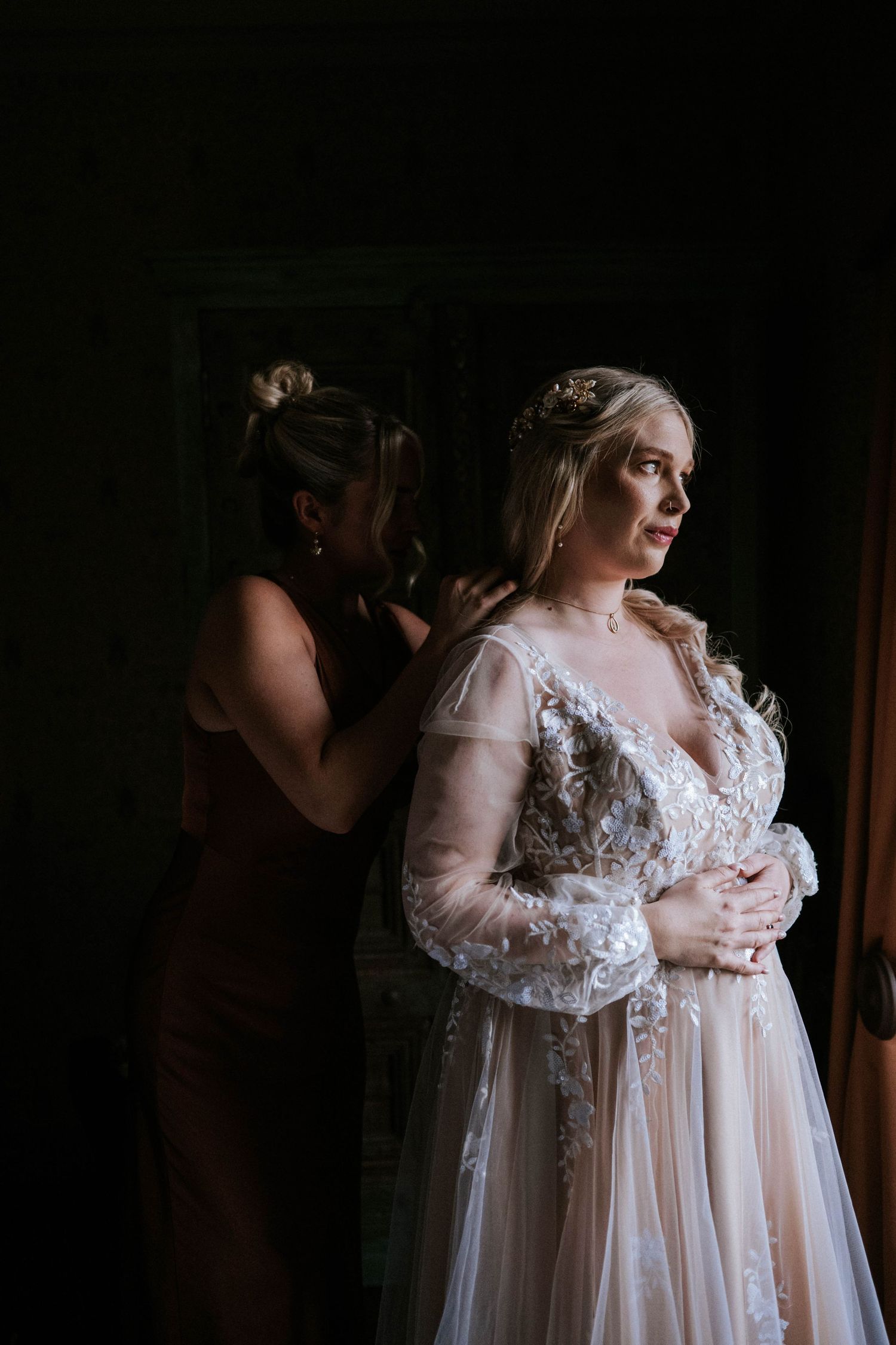 A bride in a delicate lace wedding gown with long sleeves stands in moody lighting while getting ready.