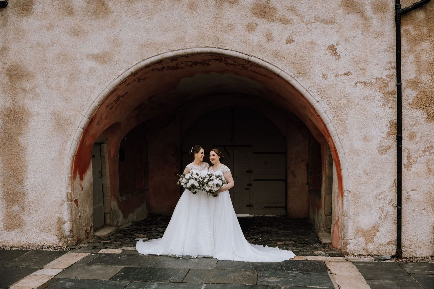 Wedding couple poses under historic terracotta archways holding white bouquets. Meldrum House Wedding Photography.
