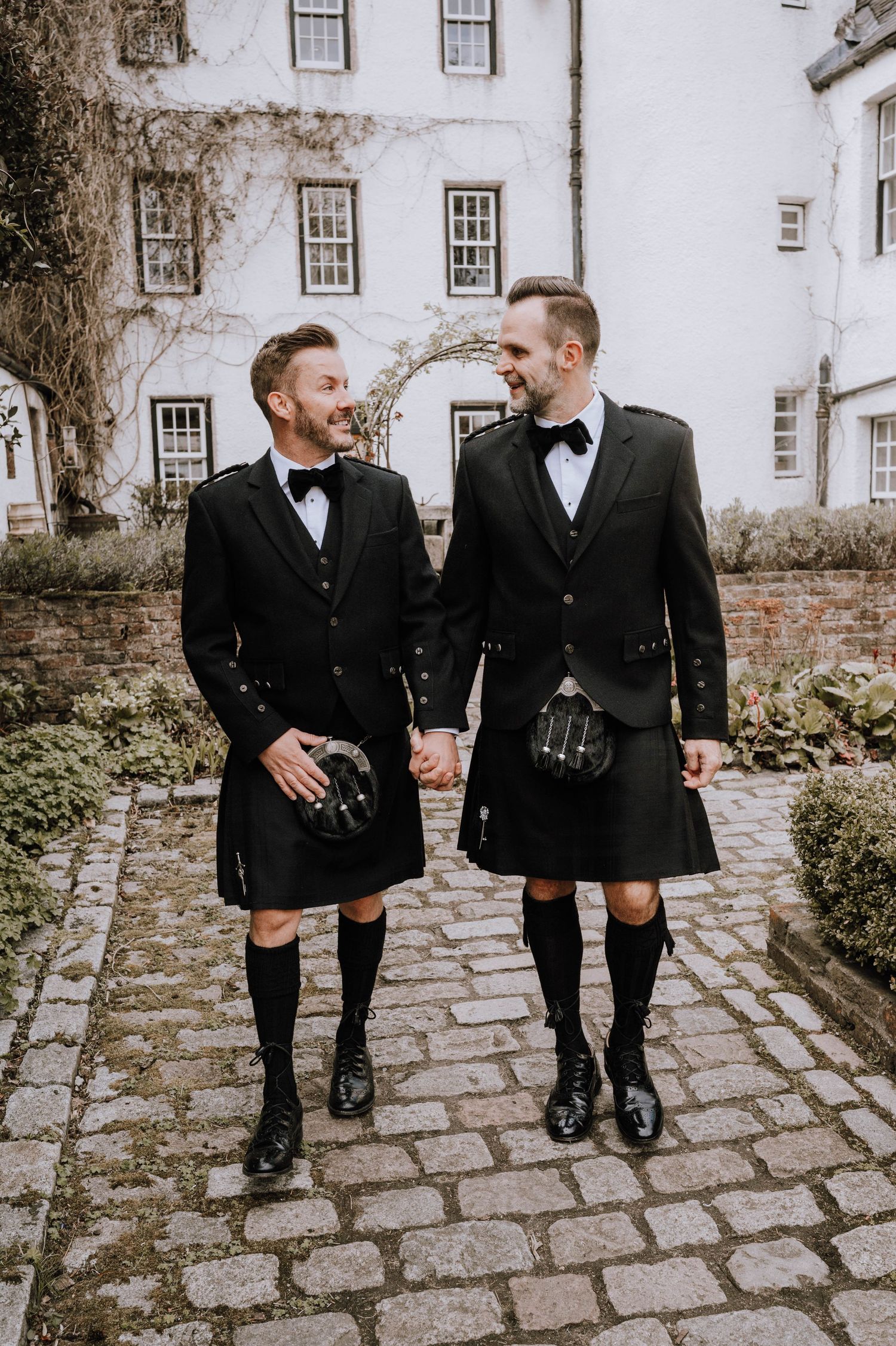 Logie House Wedding Photography. Men in traditional Scottish kilts walking along cobblestone path by historic building.