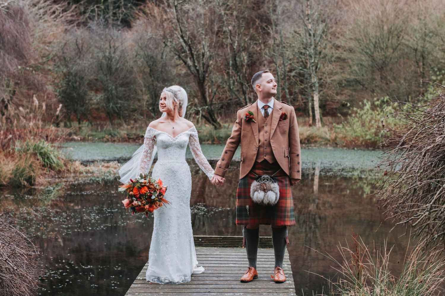 A couple walks hand in hand across a wooden bridge in a serene autumn forest setting.