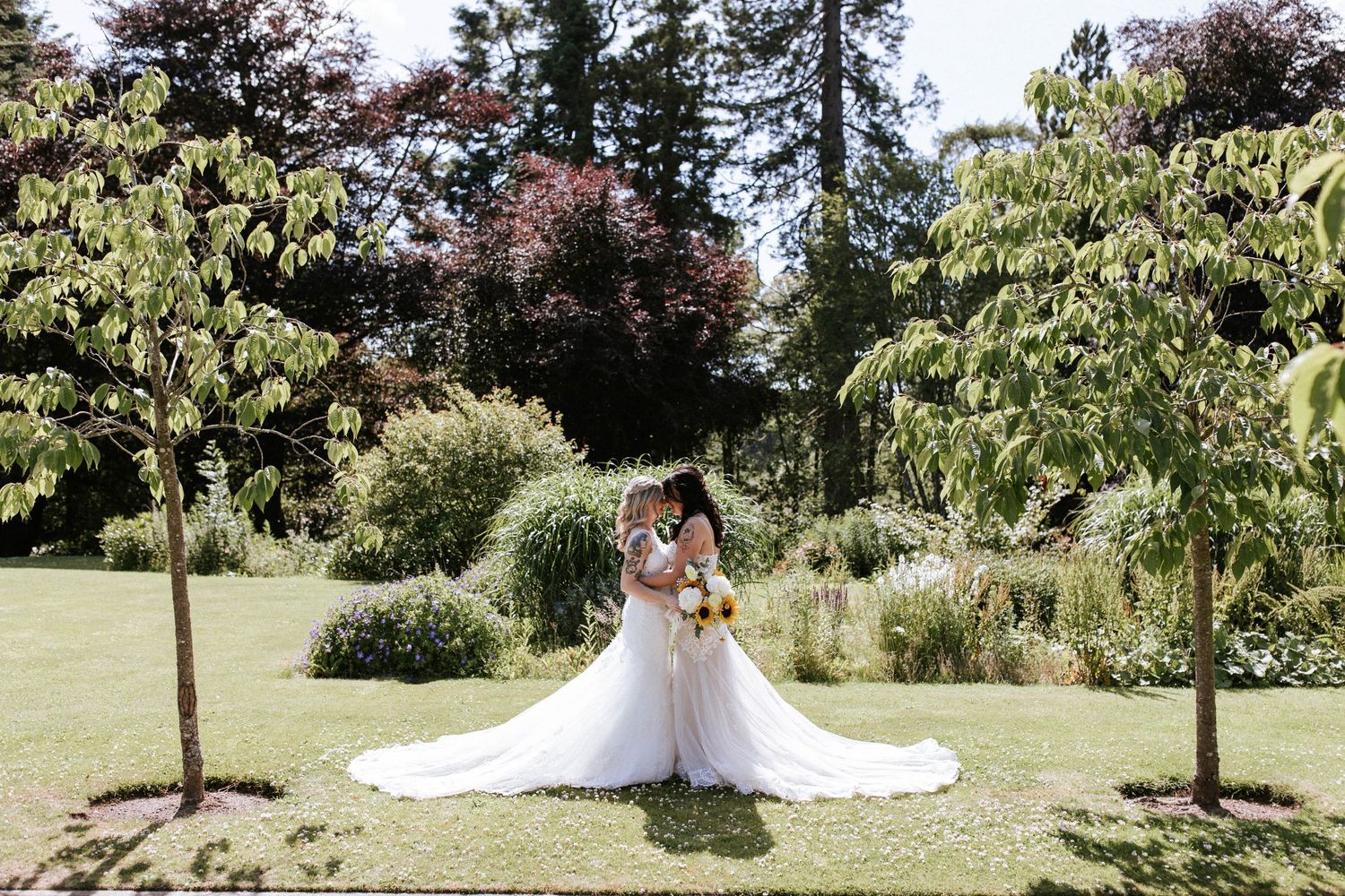 Two brides in white wedding dresses share an intimate moment in a lush garden setting.