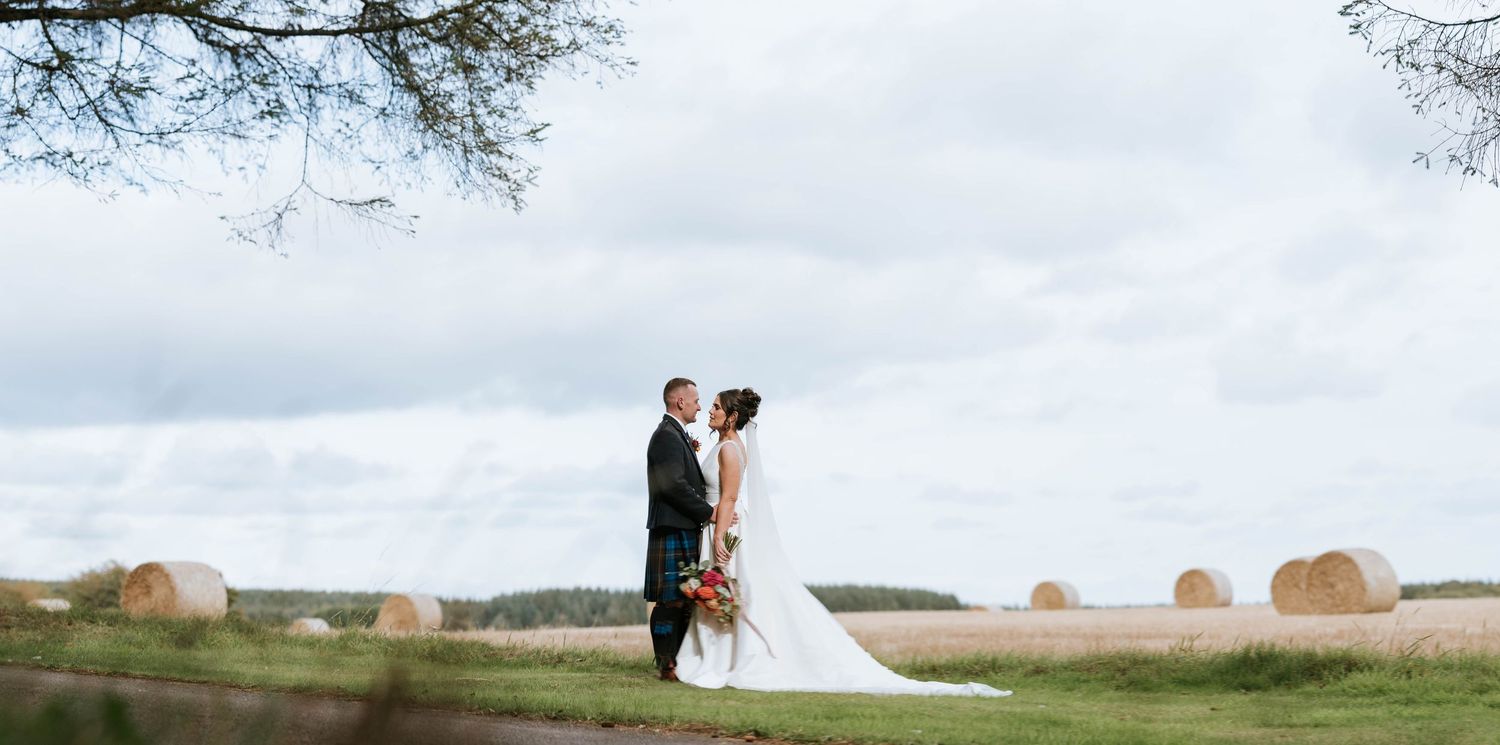 A couple stands in a field with hay bales at sunset during their Scottish wedding celebration.
