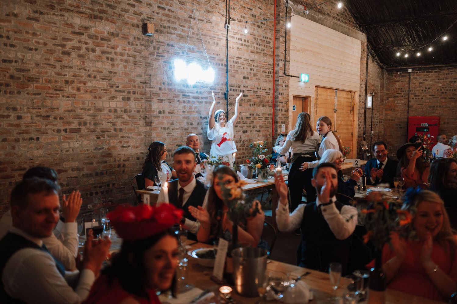 Wedding guests raise their glasses in celebration at a rustic brick venue with string lights.
