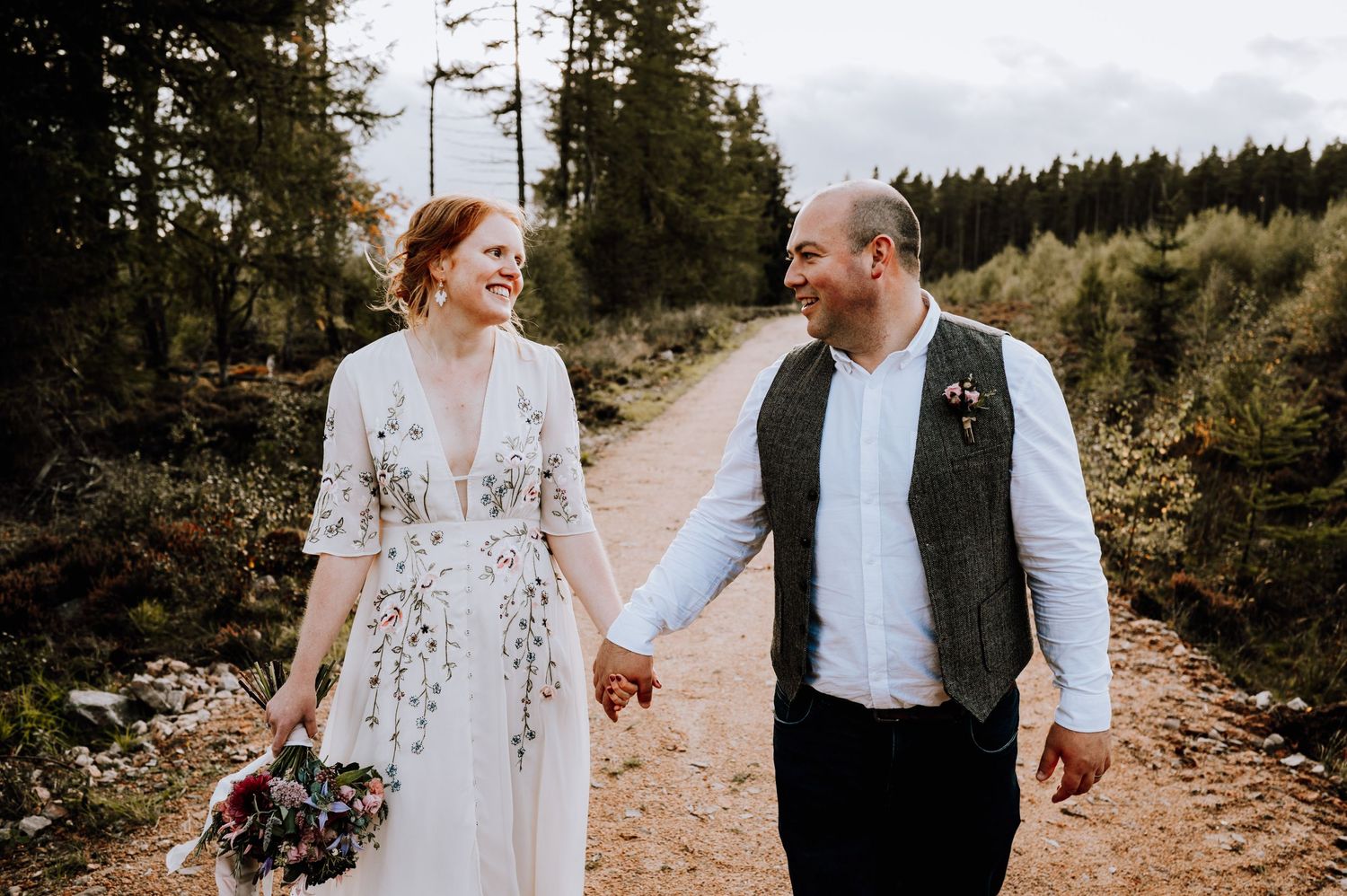 Newlyweds share joyful moment while walking hand-in-hand on forest path.