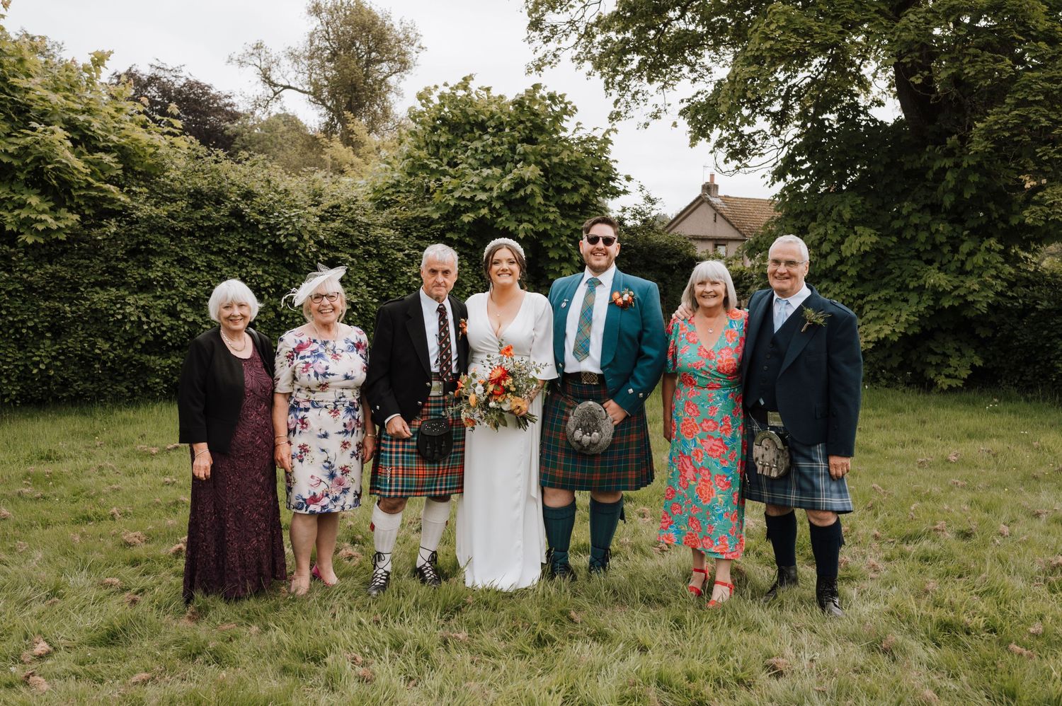 Extended family in mix of formal wear and kilts poses for wedding photo on grass.