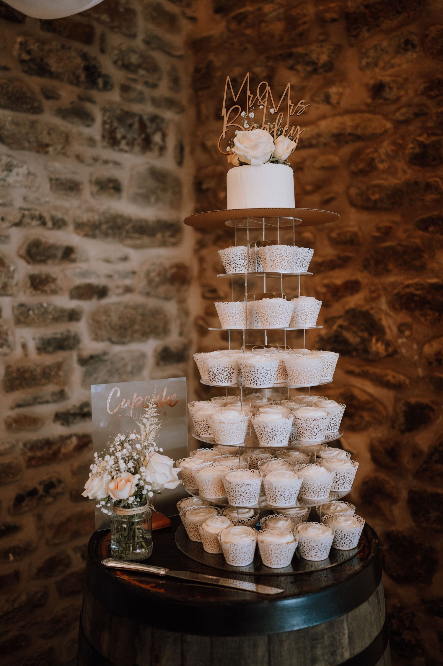 Modern wedding cake display with multiple tiers of white cupcakes and a small decorated top tier against a rustic stone wall.