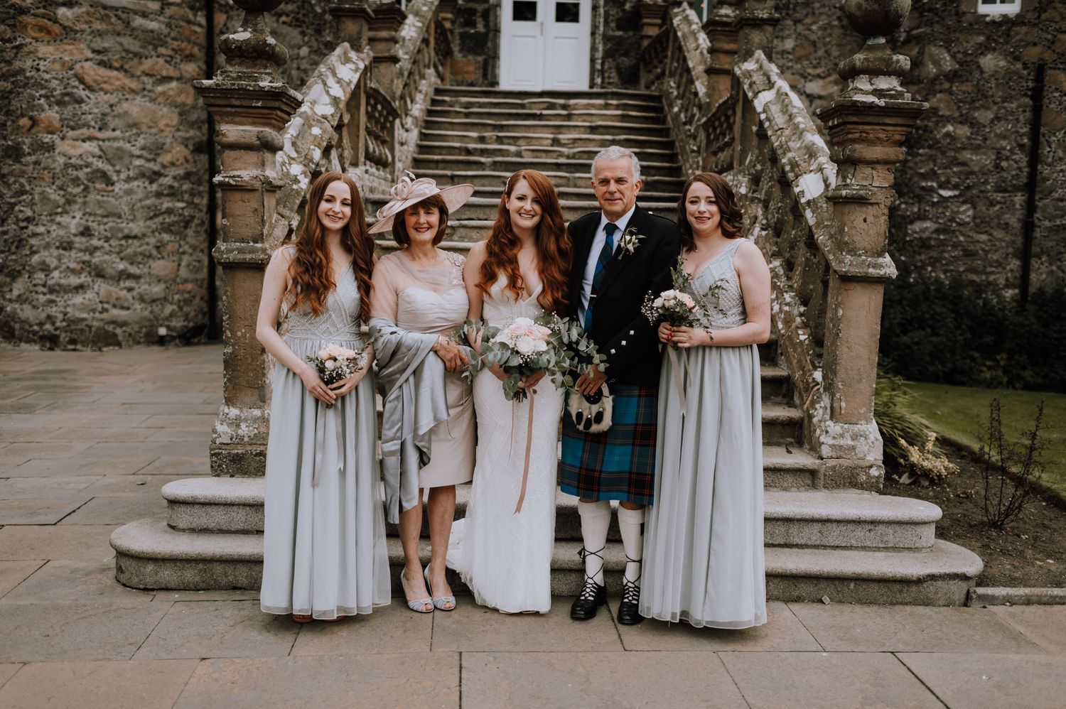 Wedding party poses on stone steps of historic building with traditional Scottish attire including kilts and formal dresses.