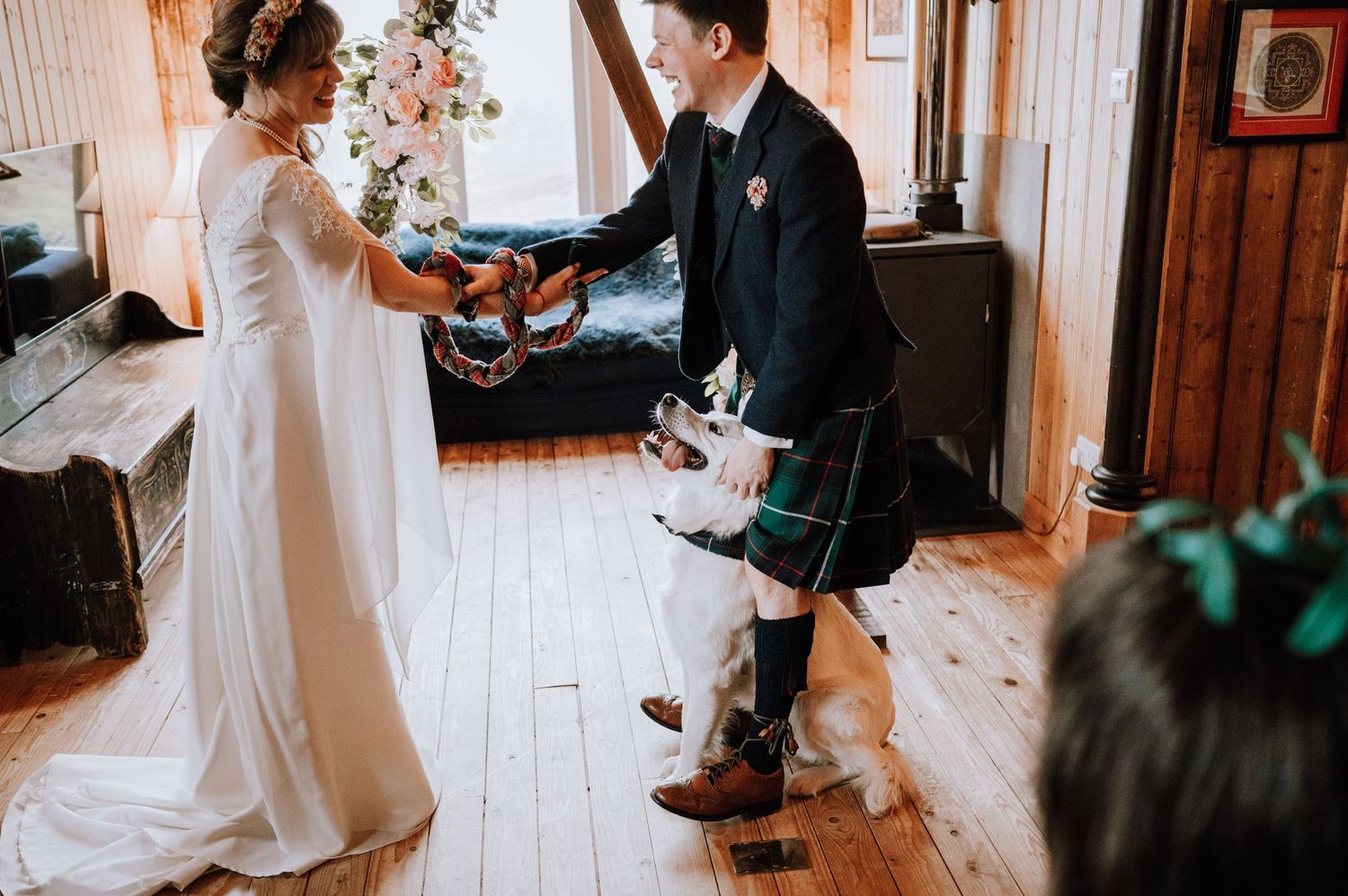 Bride in flowing white dress exchanges vows with groom in Scottish kilt at wooden chapel.