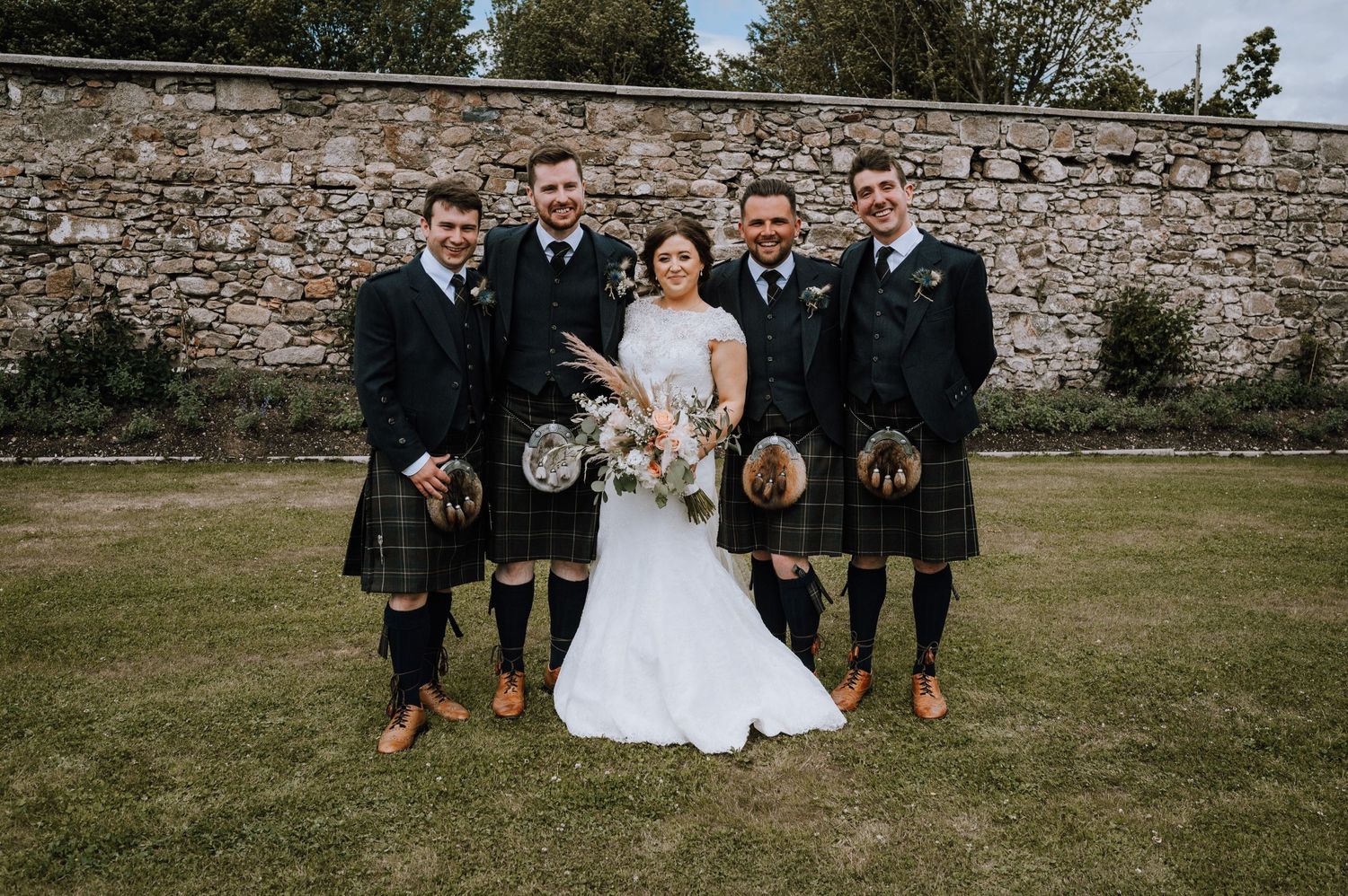 Wedding party in traditional Scottish kilts poses against stone wall.