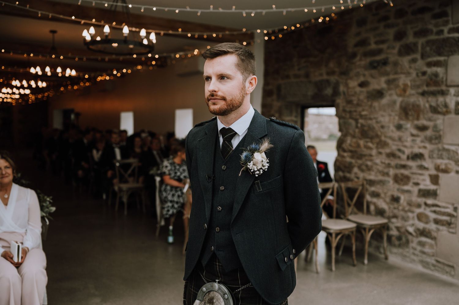 Wedding guest in traditional Scottish kilt attire stands in rustic stone-walled reception venue.