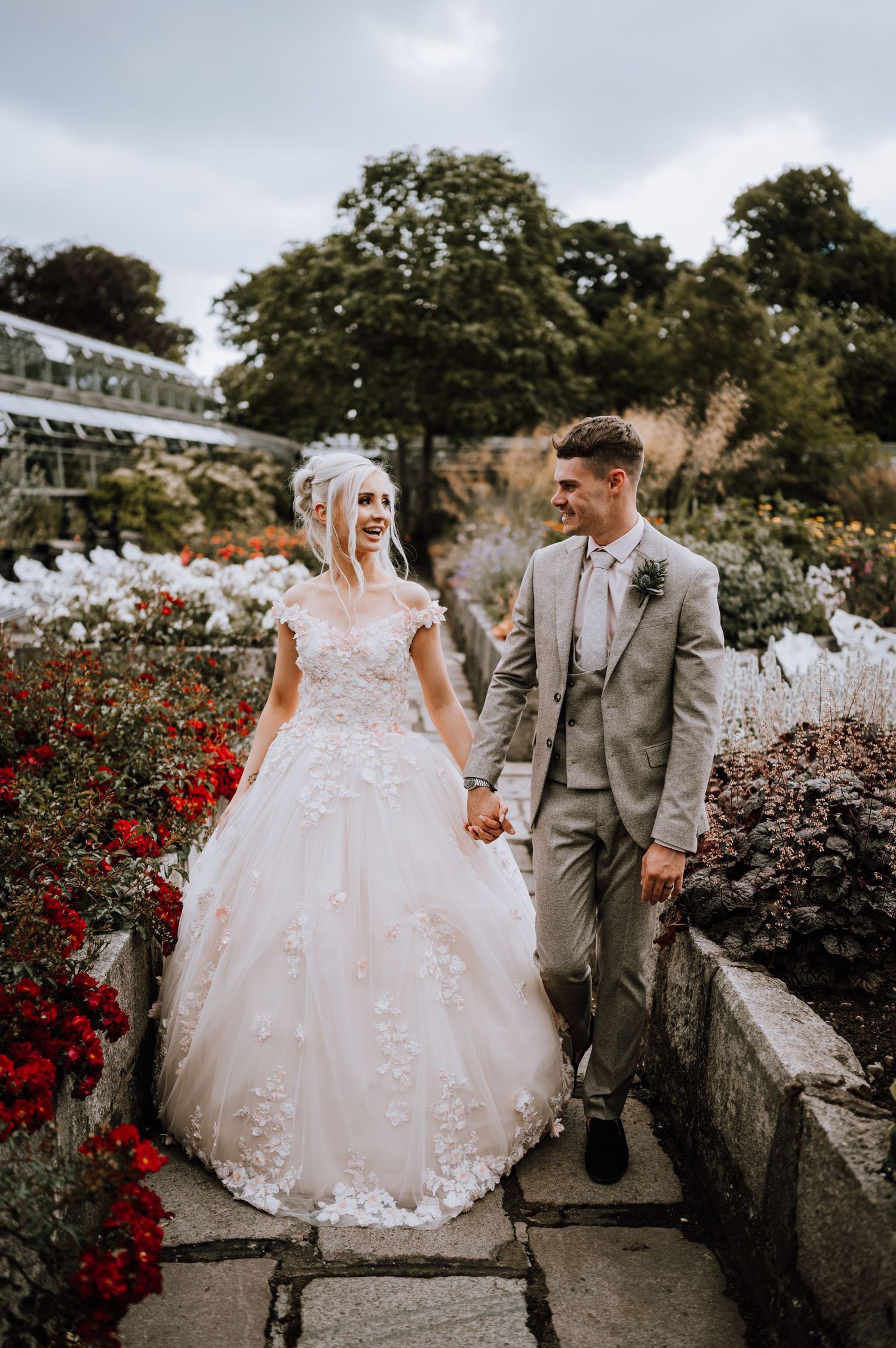A couple poses among red flowers and greenery on their wedding day.