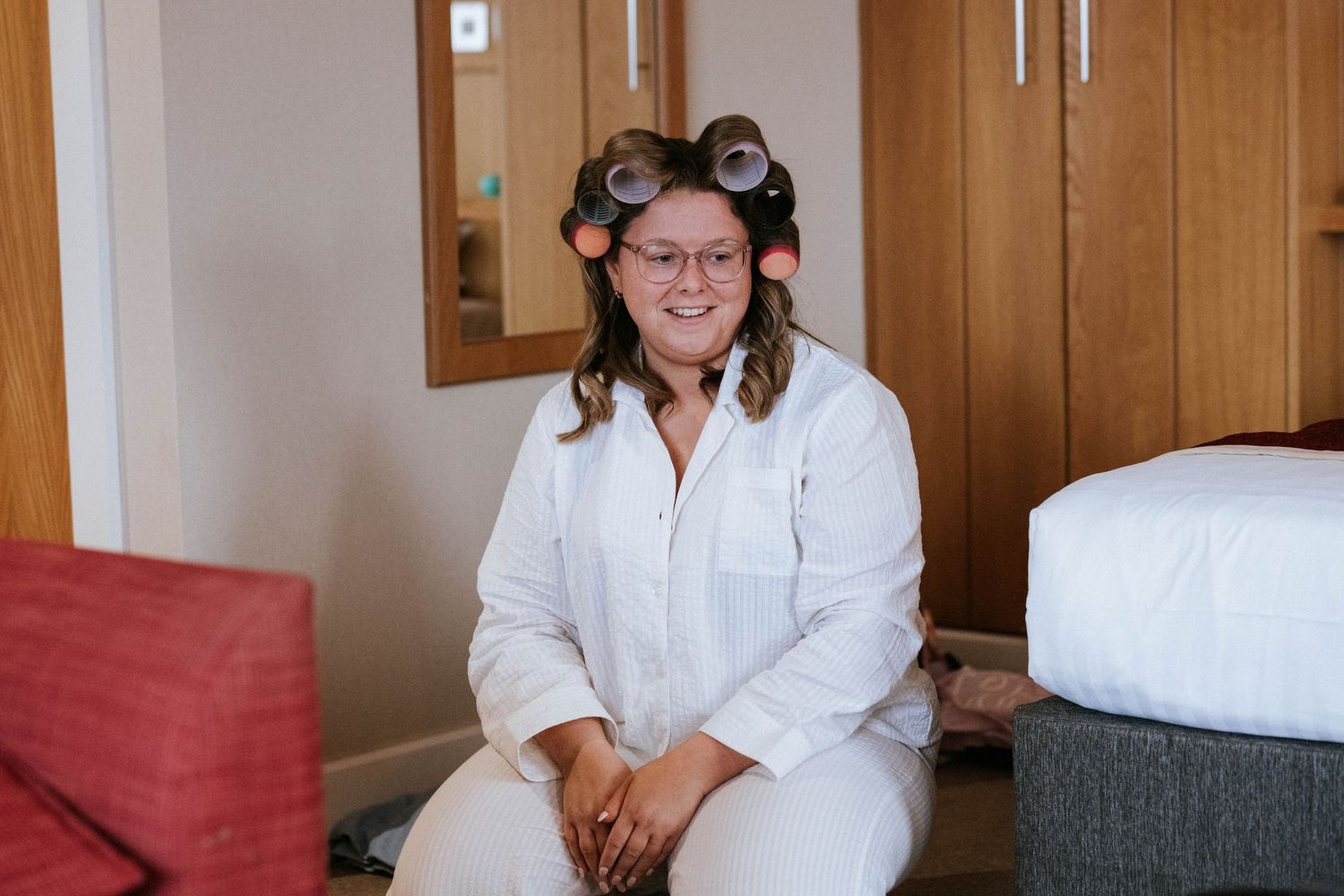 A person in white clothing and hair rollers sits smiling on a red chair during wedding preparations.
