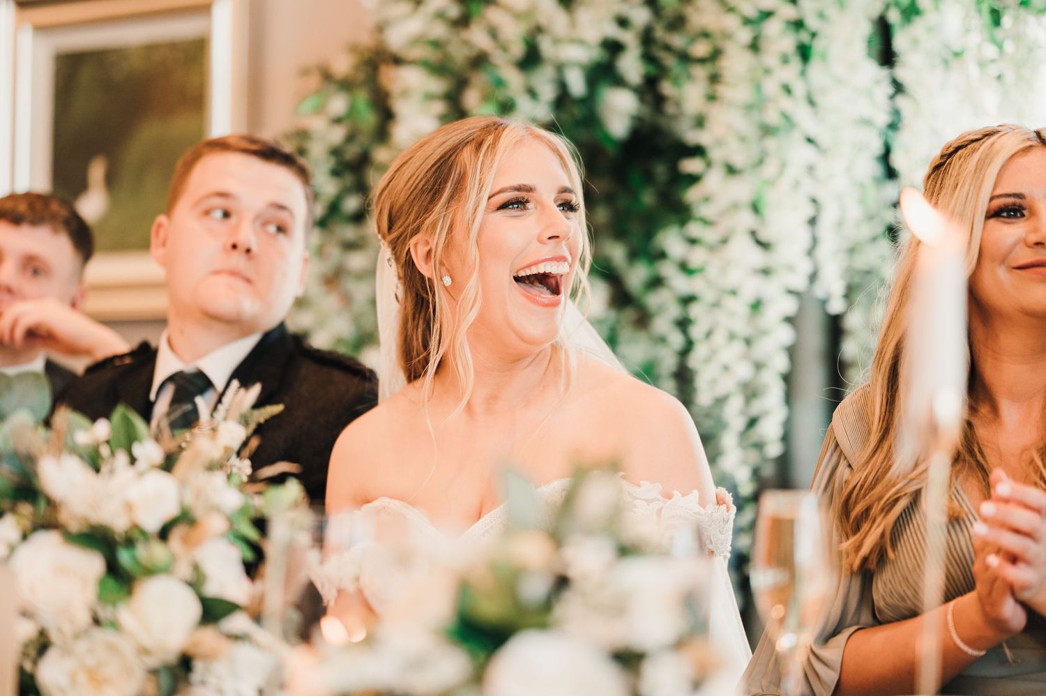 Candid reactions during wedding reception speeches with floral decorations in background.