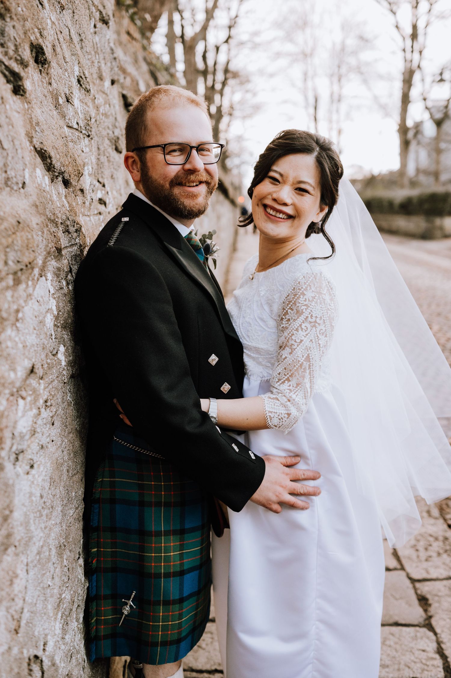 Joyful newlyweds in Scottish and traditional wedding attire share candid moment against stone wall.