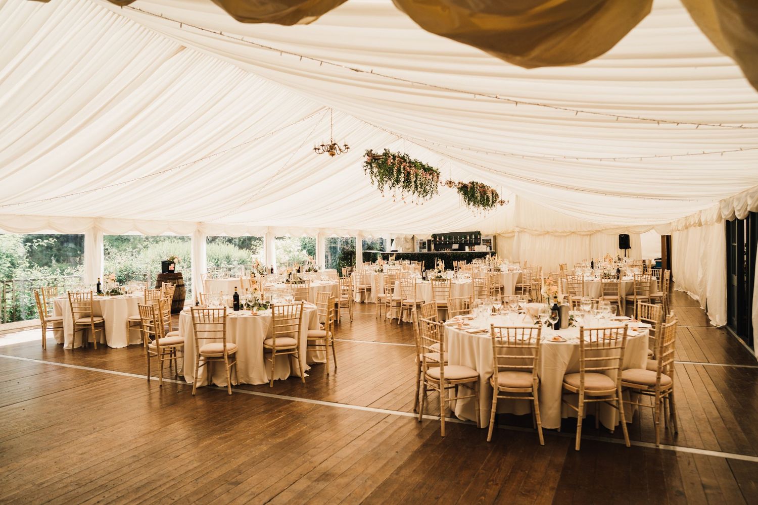 Elegant wedding reception setup in white marquee tent with wooden floor and gold chiavari chairs.