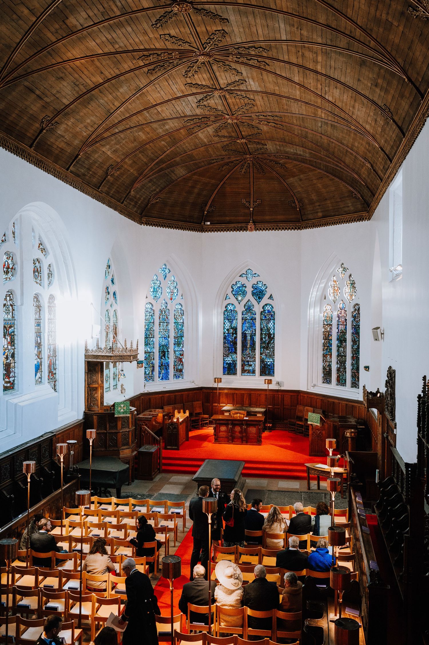 Church interior with wooden vaulted ceiling, stained glass windows, and red carpeted altar area.