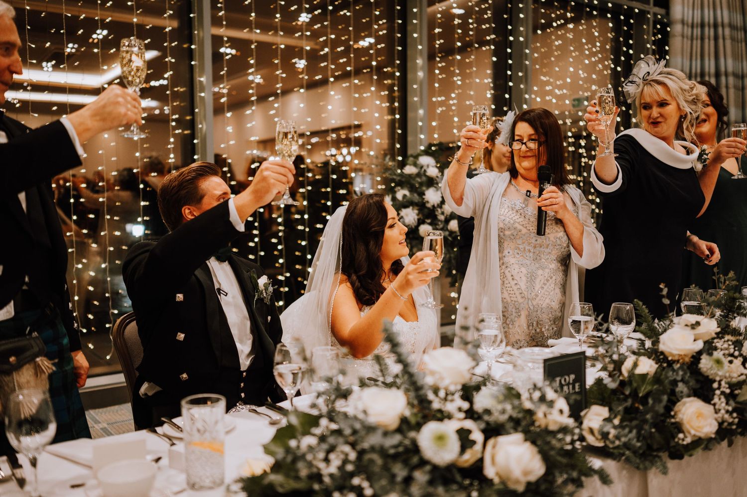 Wedding party raises glasses for a toast against a backdrop of string lights and floral centerpieces.