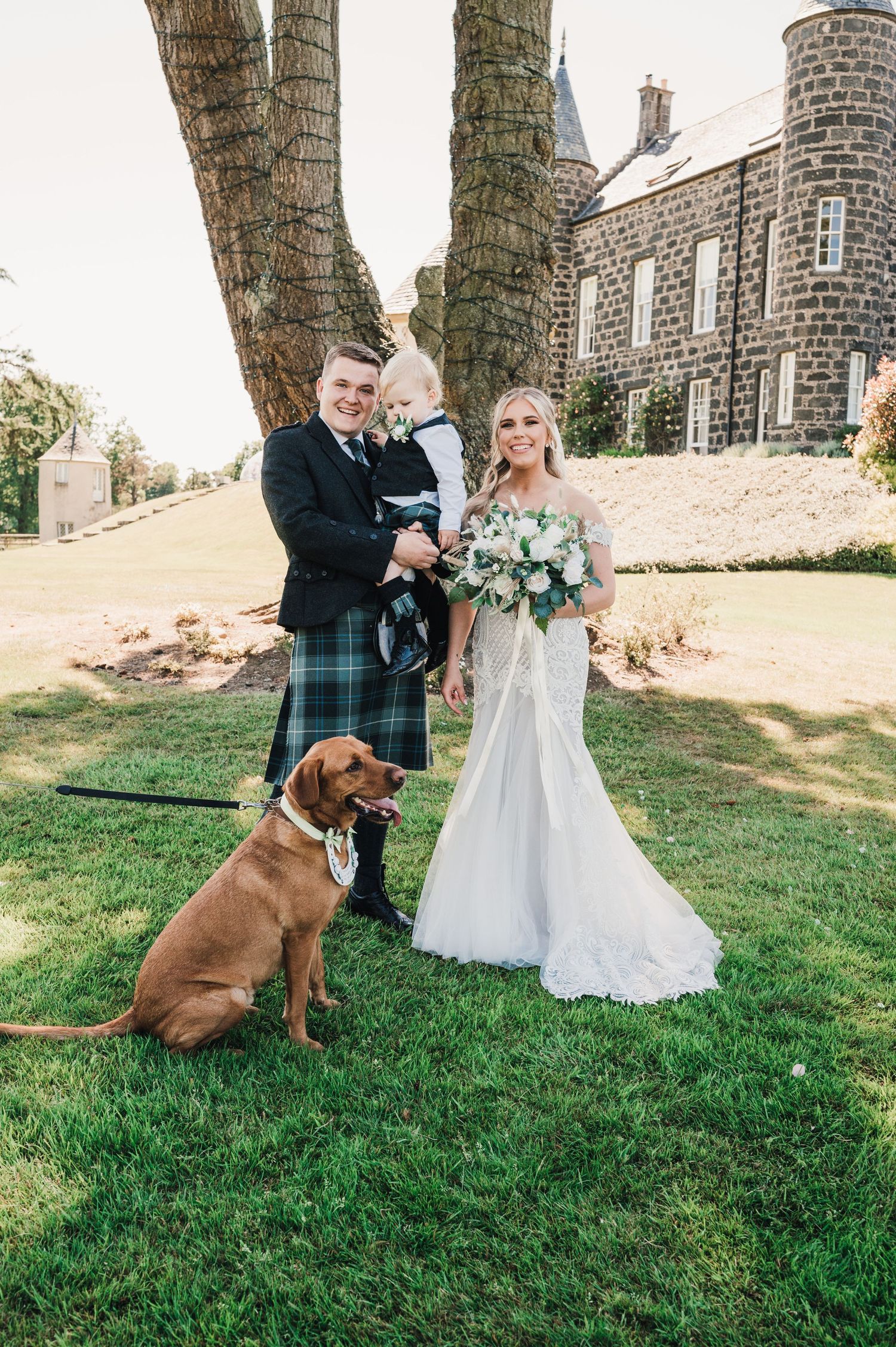 A wedding portrait featuring a couple in Scottish attire with their brown dog on a lawn.