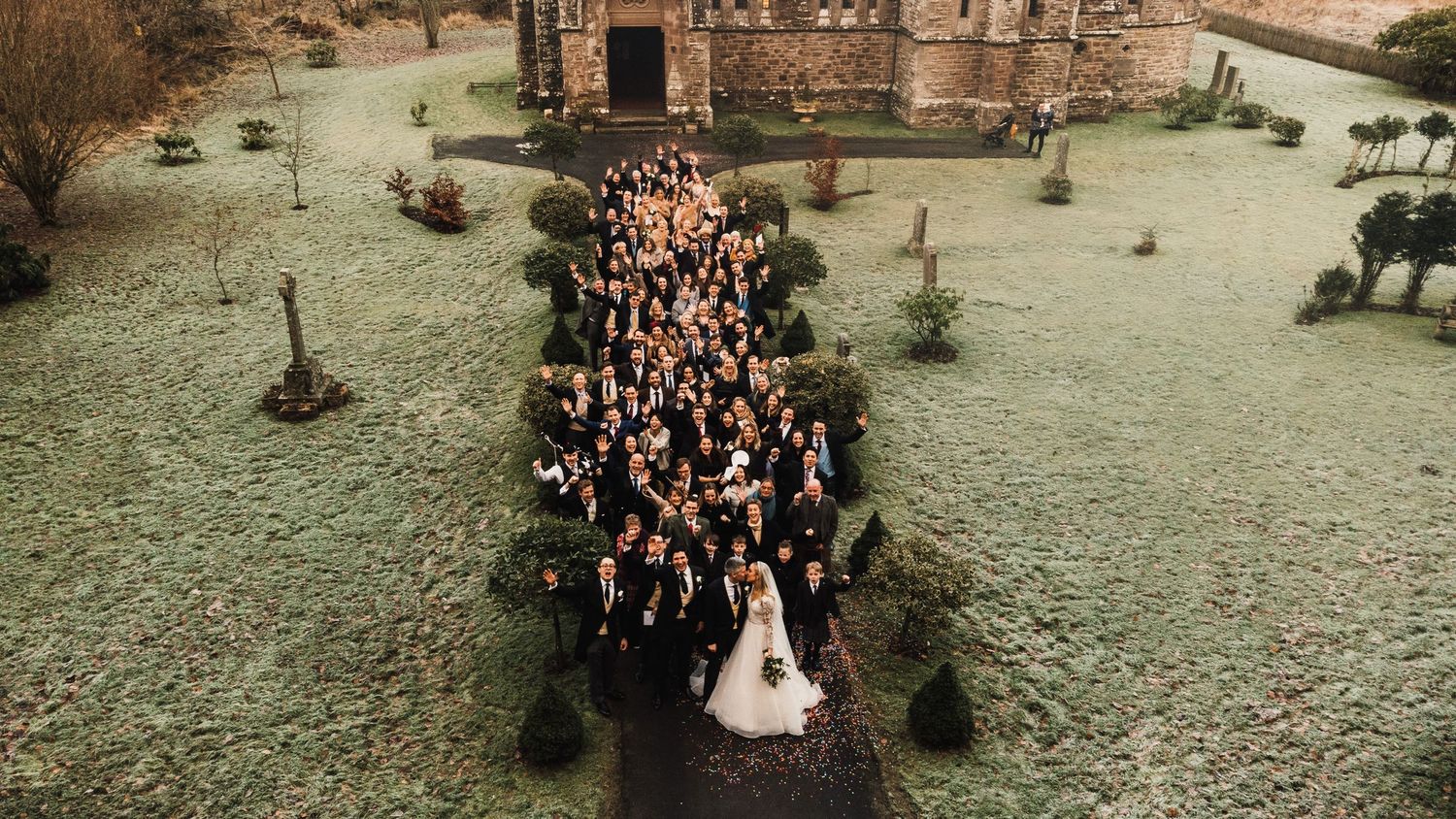 Aerial view of wedding guests gathered outside a historic stone church on a grassy lawn.
