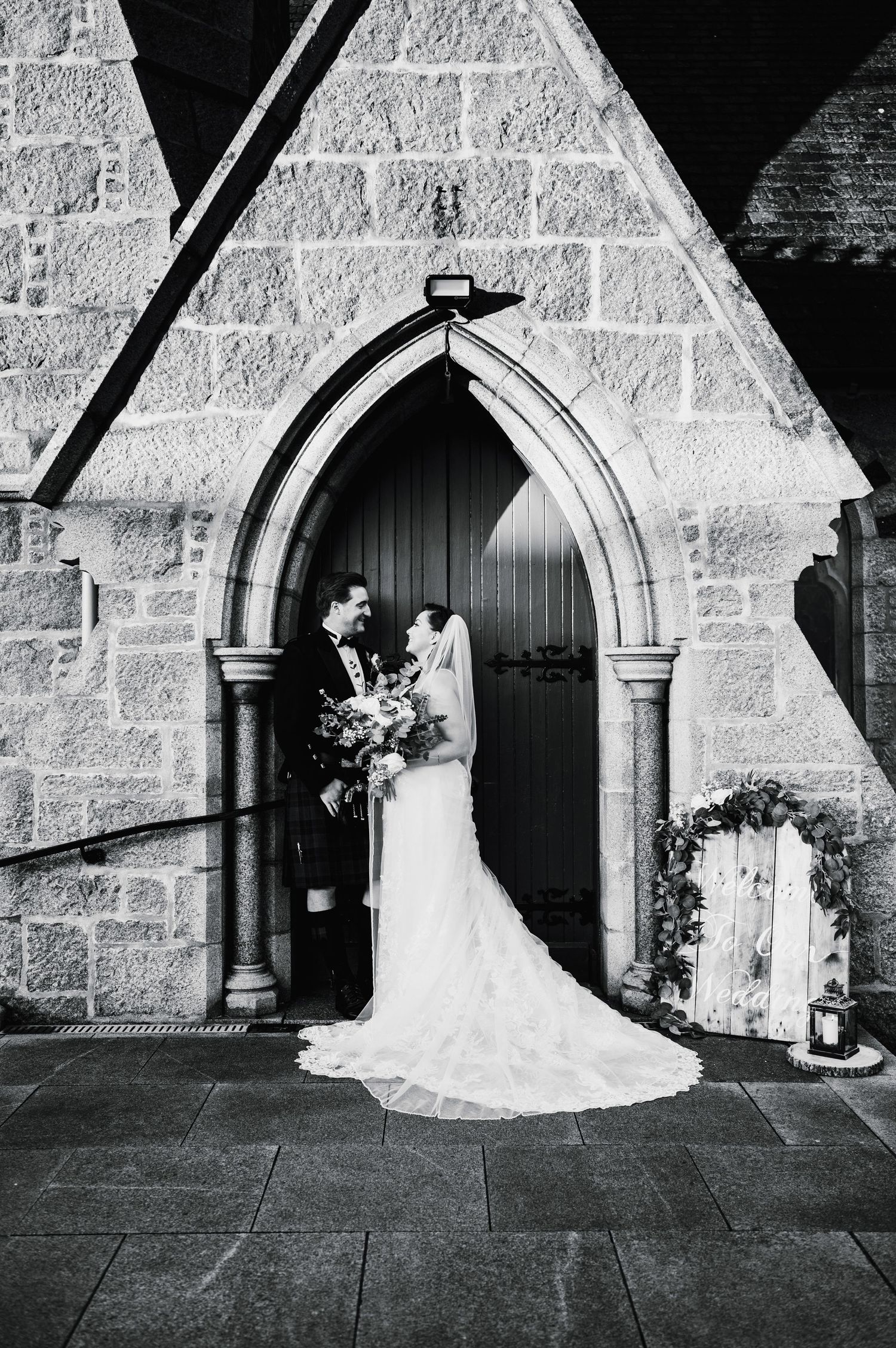 Black and white portrait of couple standing in gothic church doorway with trailing wedding dress.