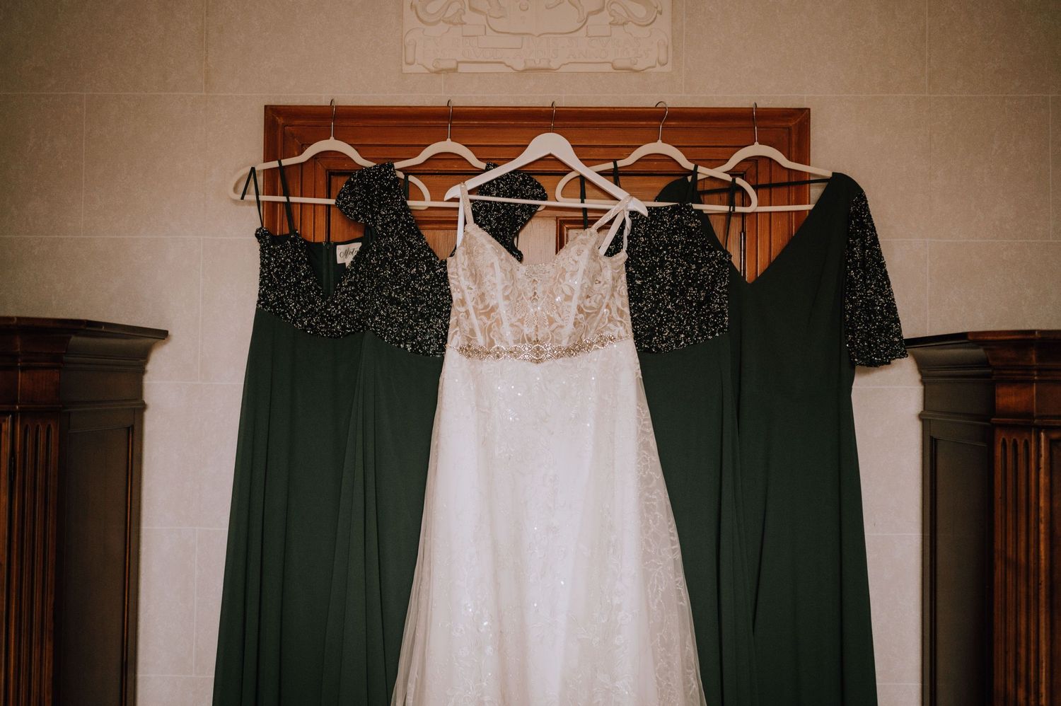 A white wedding dress hangs between two dark green bridesmaid dresses on wooden display hooks.