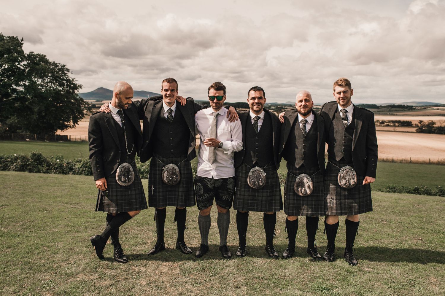 Wedding party in traditional Scottish kilts poses on hilltop with scenic landscape background.