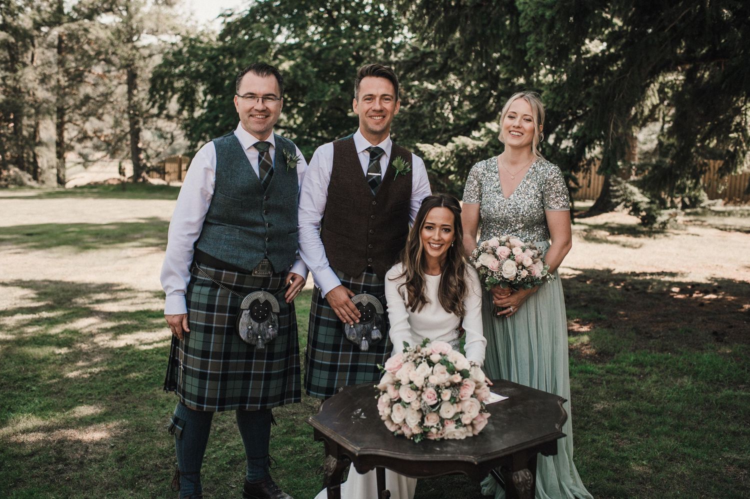 Wedding party poses outdoors with Scottish kilts and formal attire while displaying a beautiful floral bouquet.