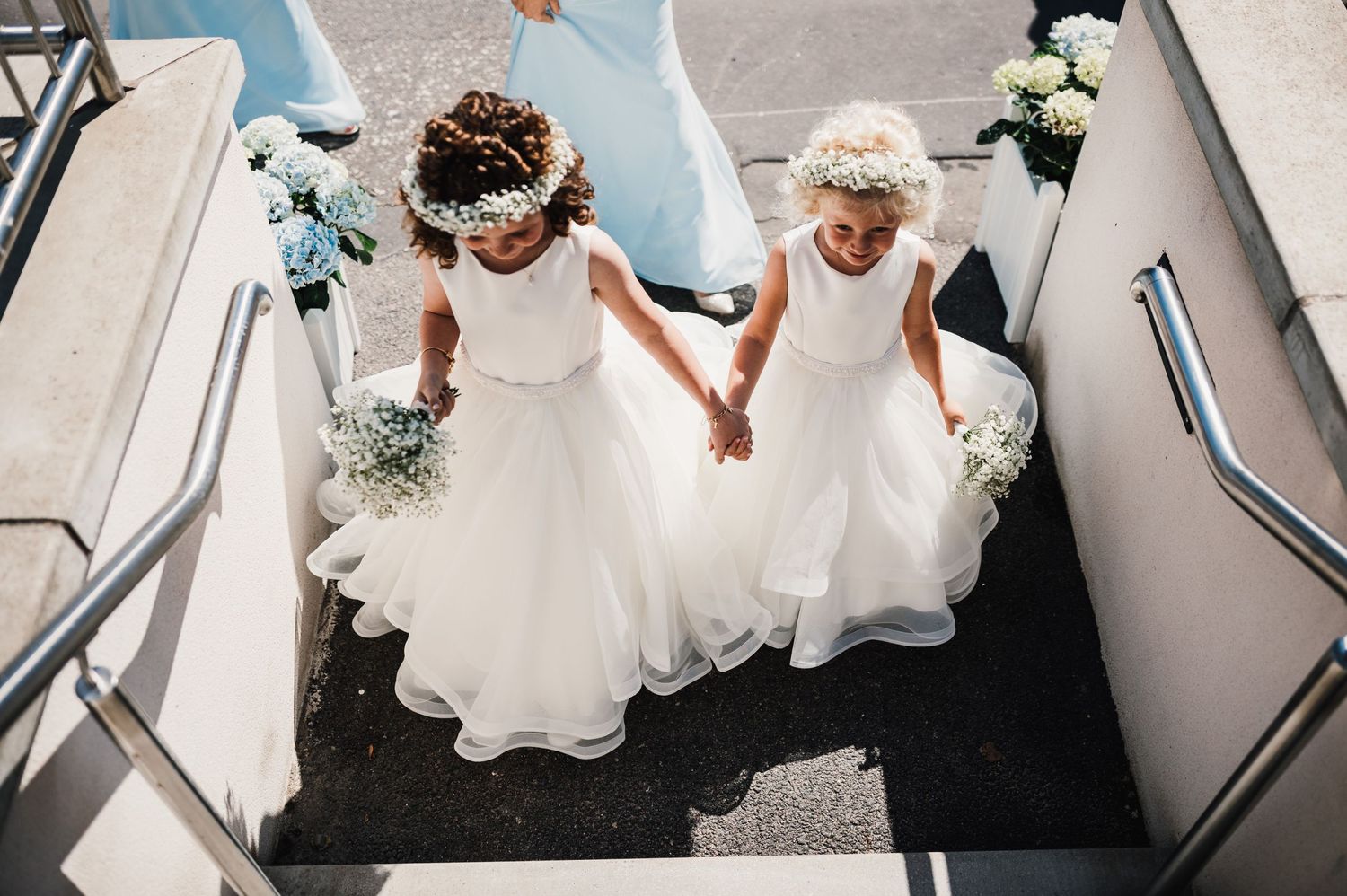 Two flower girls in white dresses holding hands while walking down church steps.