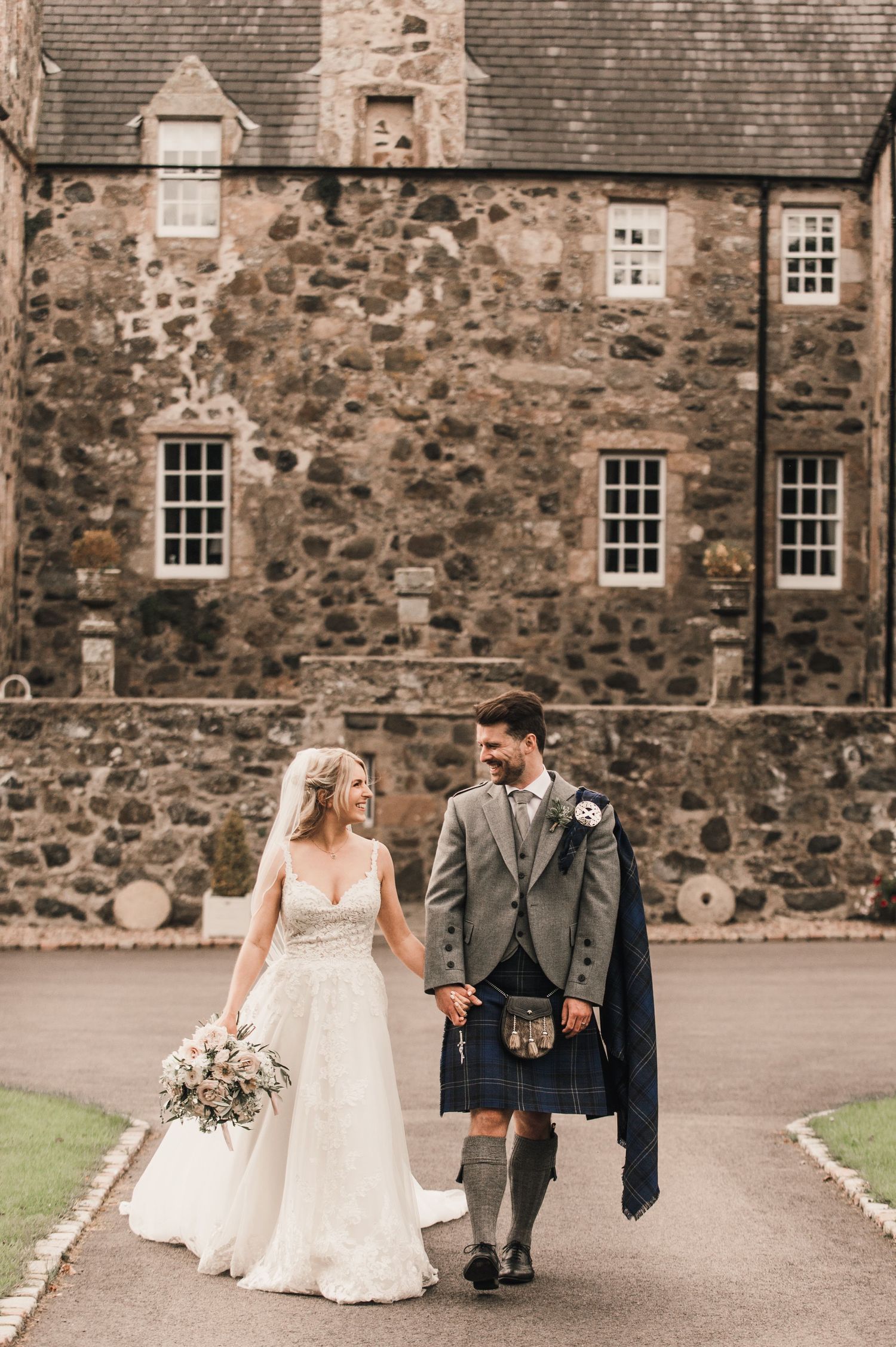 Couple in wedding attire including traditional Scottish kilt walking outside historic stone castle building.