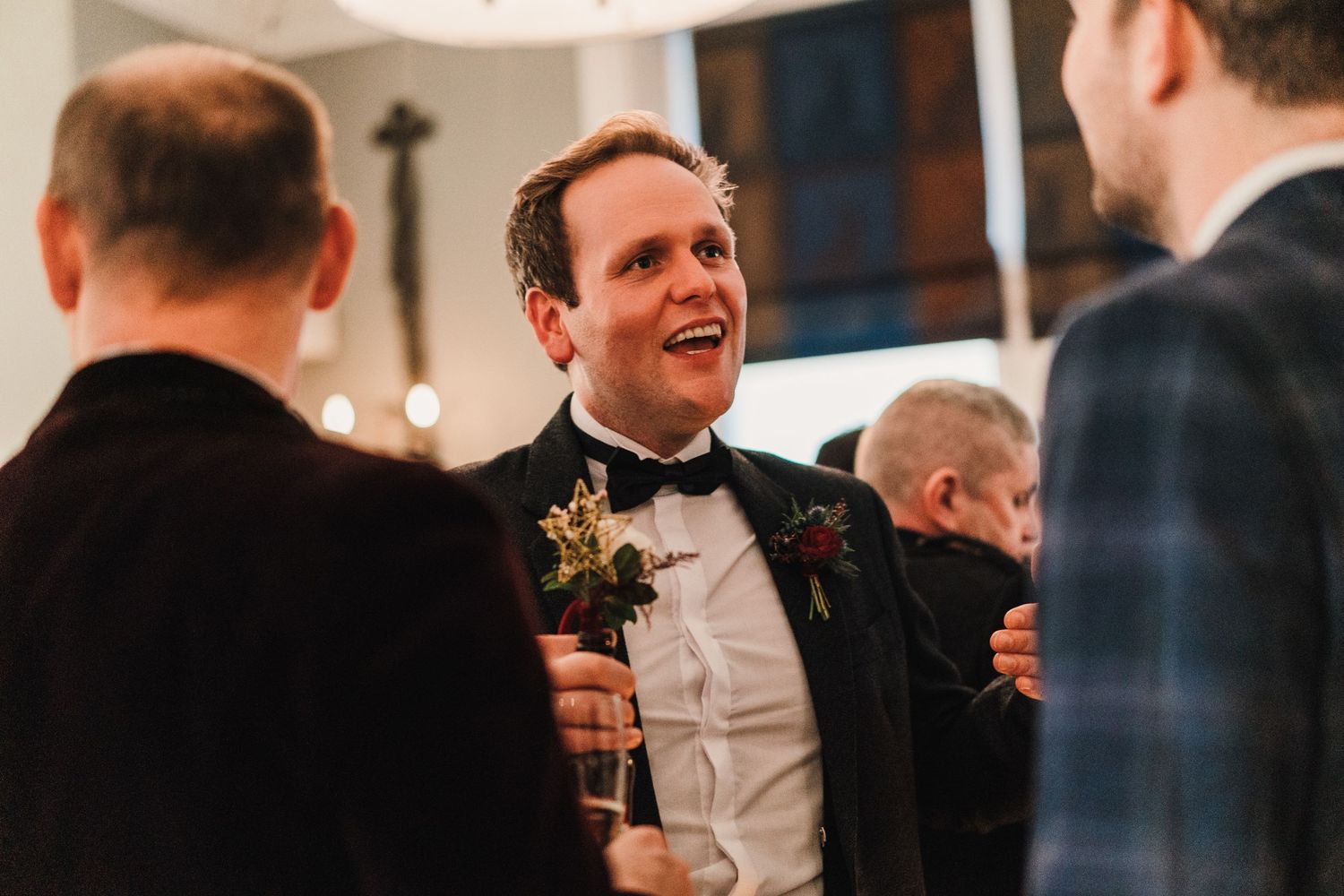 A candid moment at a formal wedding reception shows guests conversing and laughing in black tie attire.