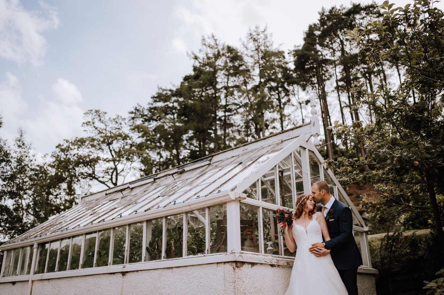 Elegant wedding portrait in front of white conservatory greenhouse with towering pine trees.