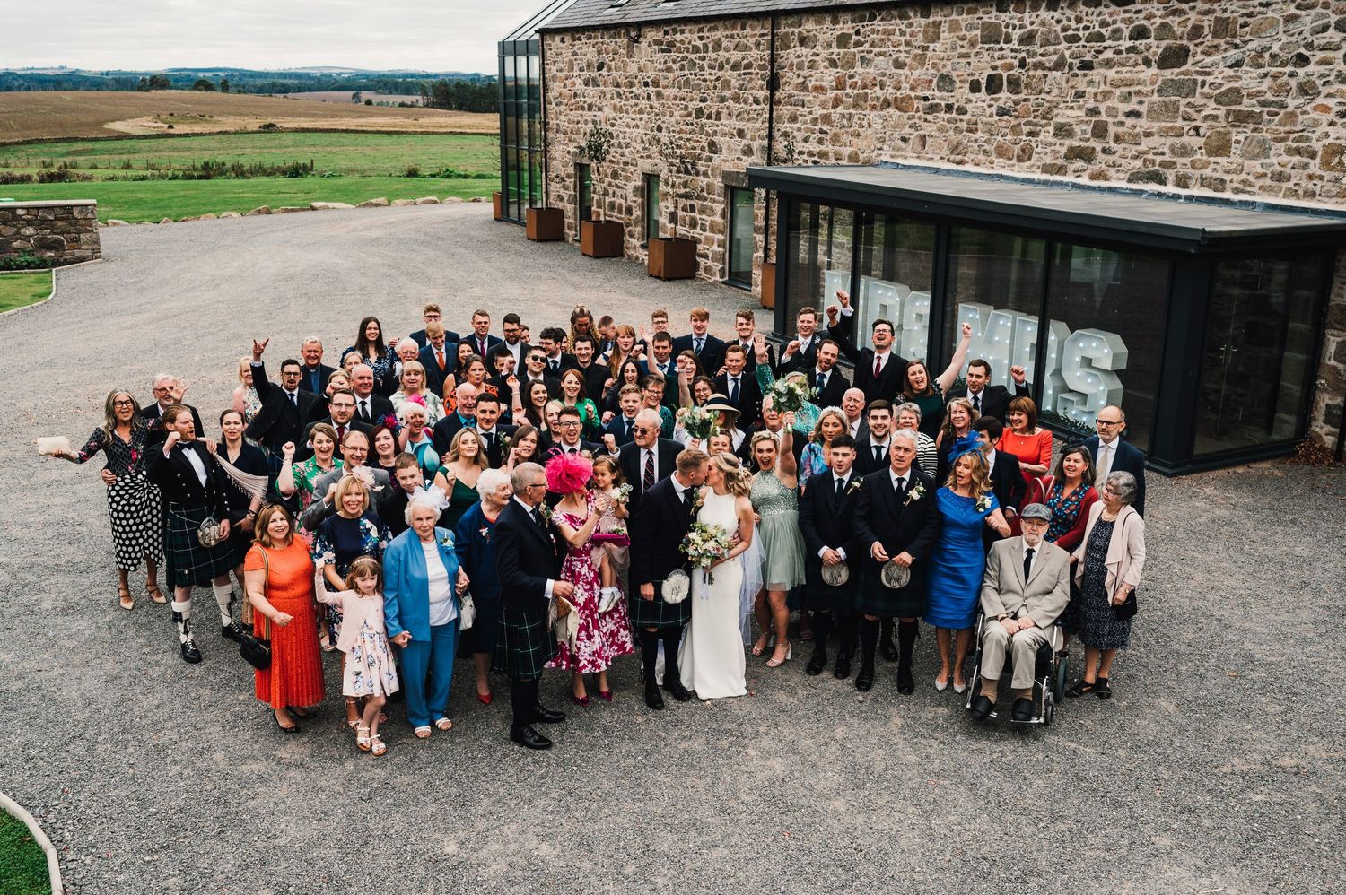 Aerial view of large wedding group photo with guests gathered outside stone building venue.