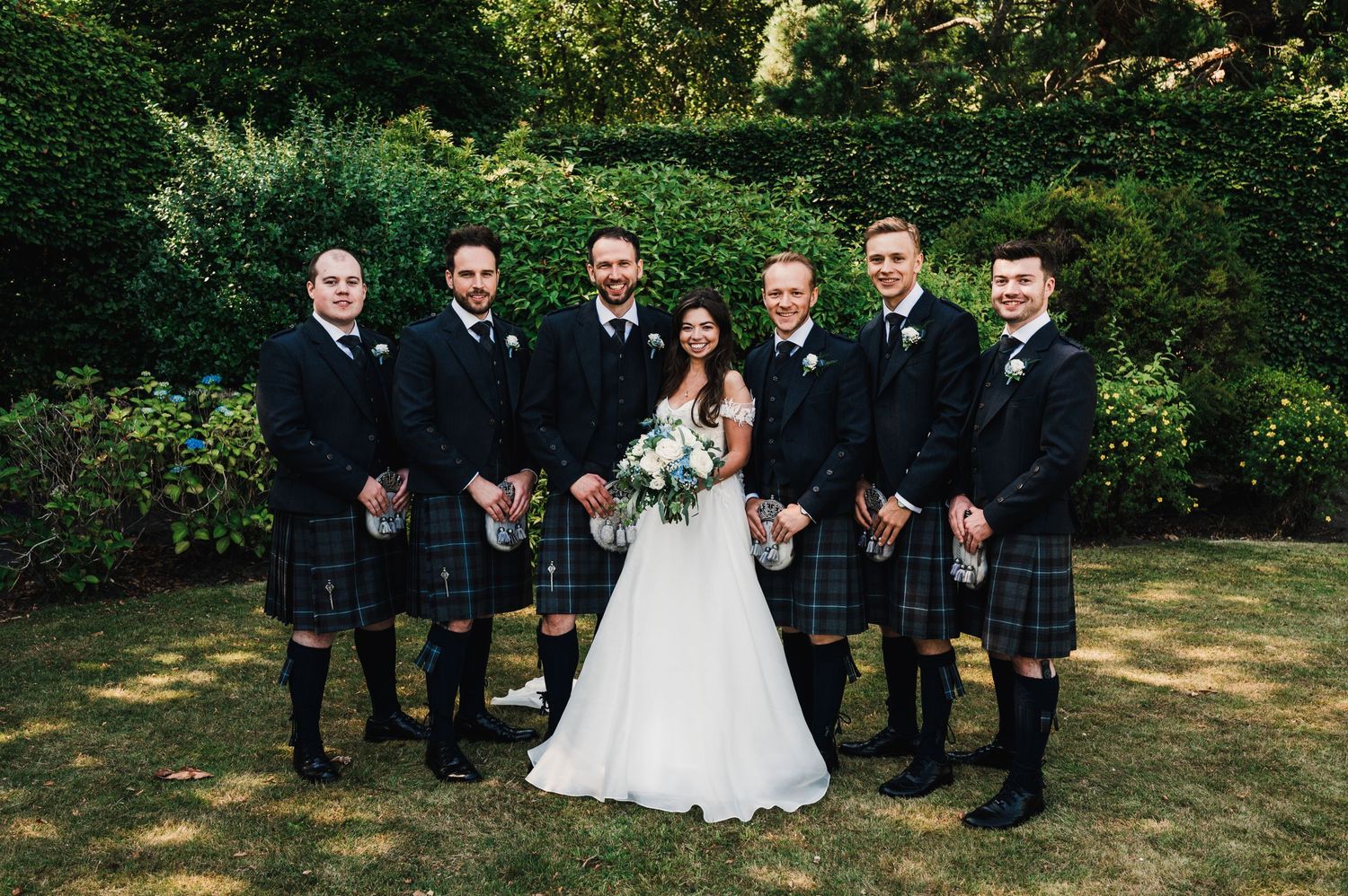 Groomsmen in traditional Scottish kilts pose with couple on garden lawn.
