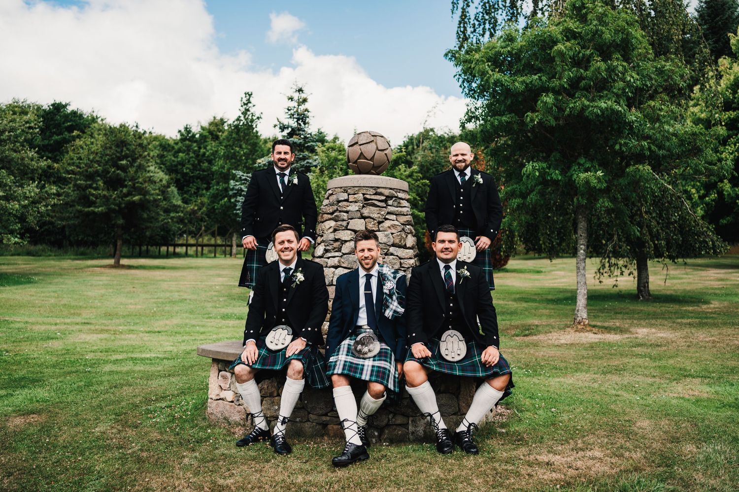 A group in traditional Scottish kilts pose together on a stone structure in a green garden setting.