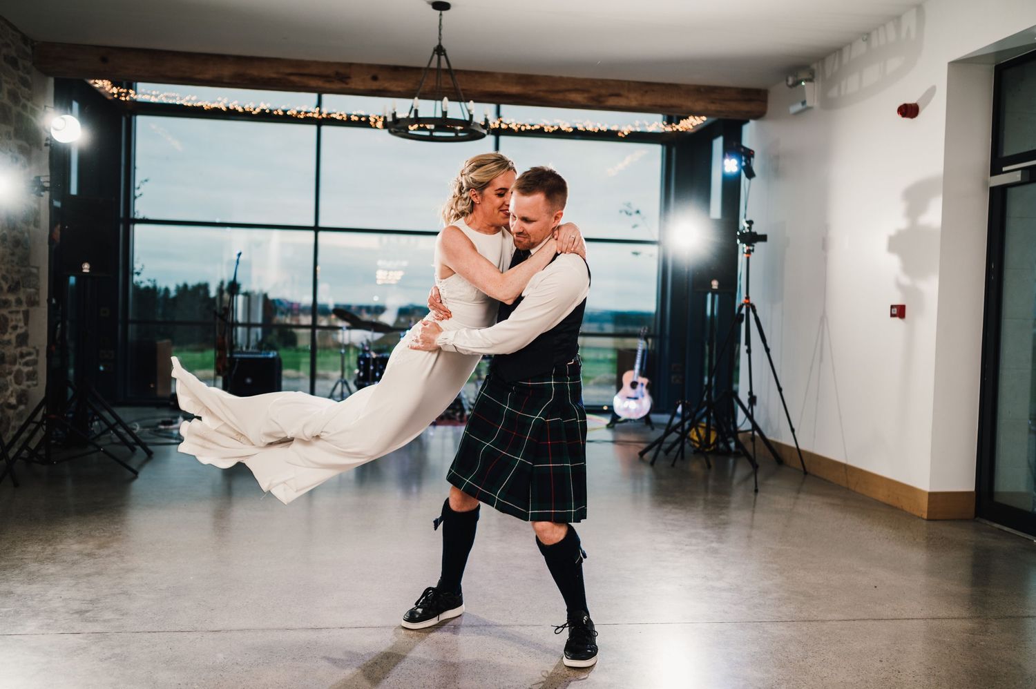 Groom in traditional kilt spins bride during wedding dance in front of large windows.