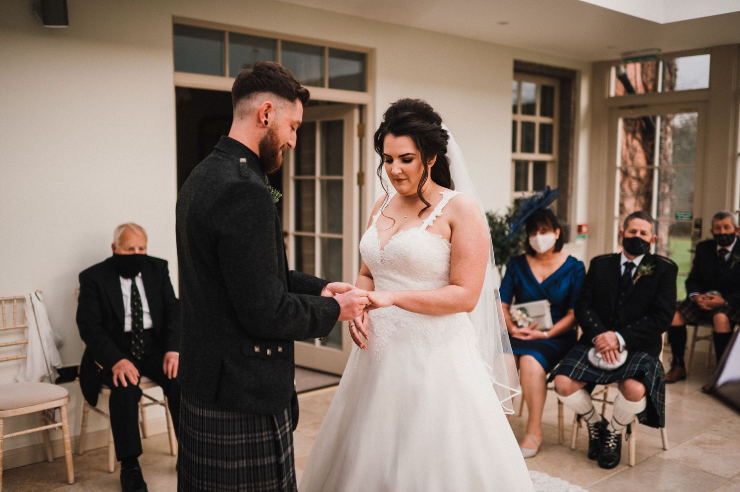 A couple exchanges rings during a Scottish-themed wedding ceremony with seated guests.