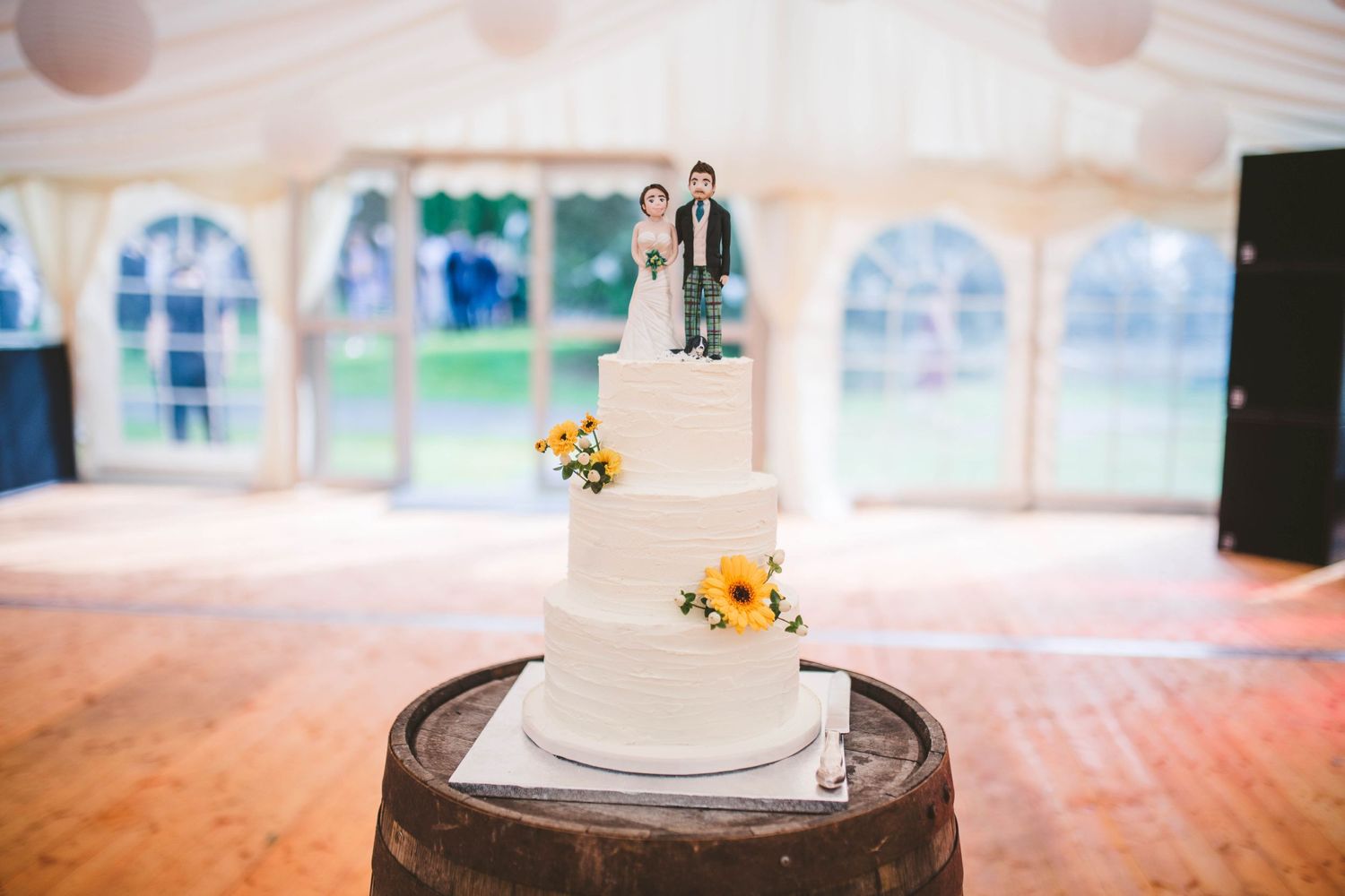 Simple white wedding cake with sunflower decorations displayed on a rustic wooden barrel.