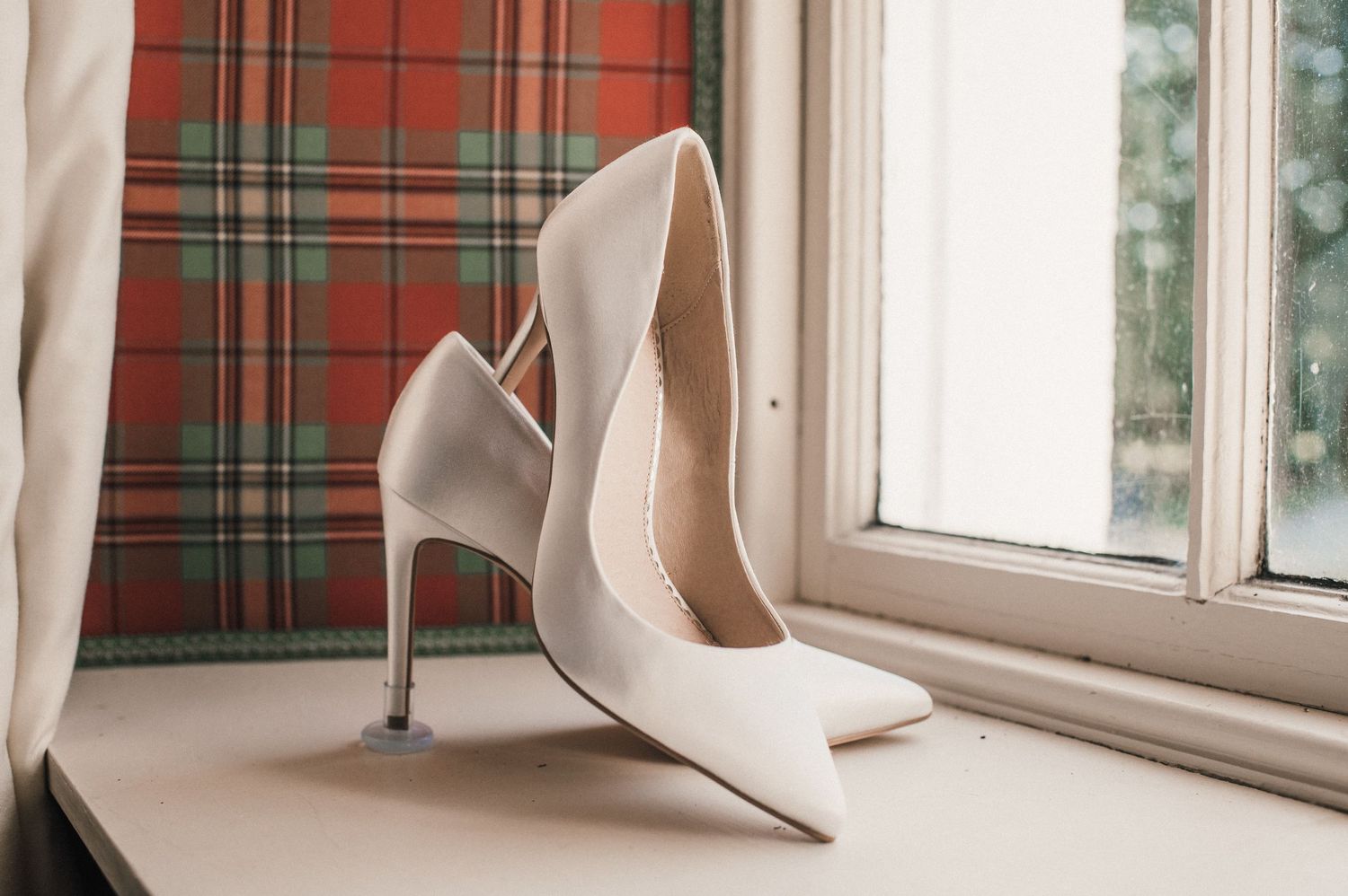 Elegant white stiletto heels displayed on a windowsill with plaid curtains.