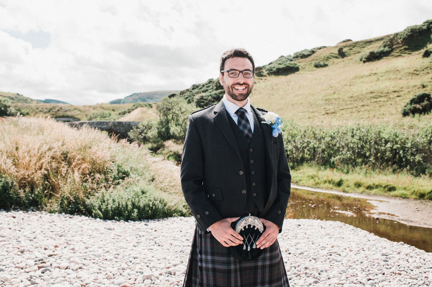 A groom in traditional Scottish formal wear stands on a rocky path with hills in the background.
