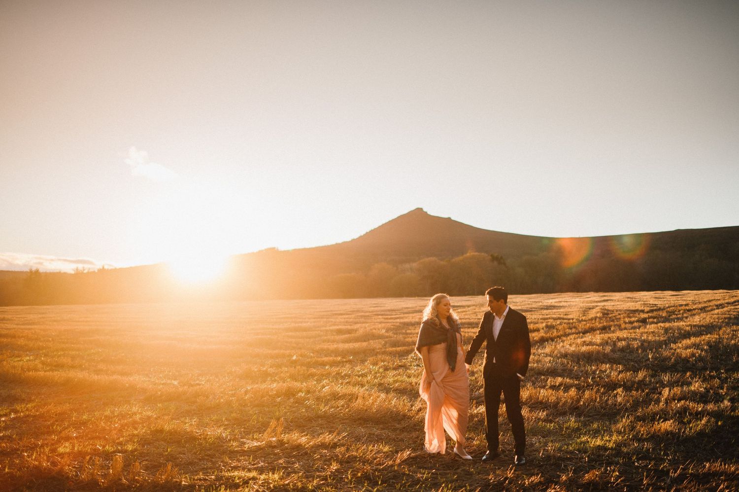 A couple walks hand in hand across a sunlit field with mountains in the distance.