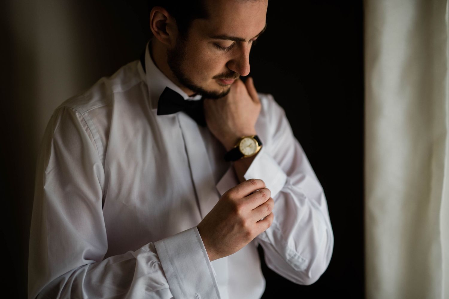 Person in white dress shirt and bow tie adjusting their watch in moody lighting.