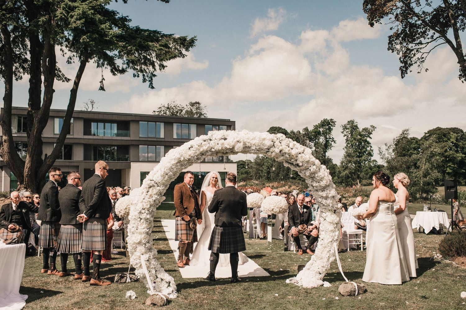 Outdoor wedding ceremony setup with white floral arch and guests in traditional Scottish dress.