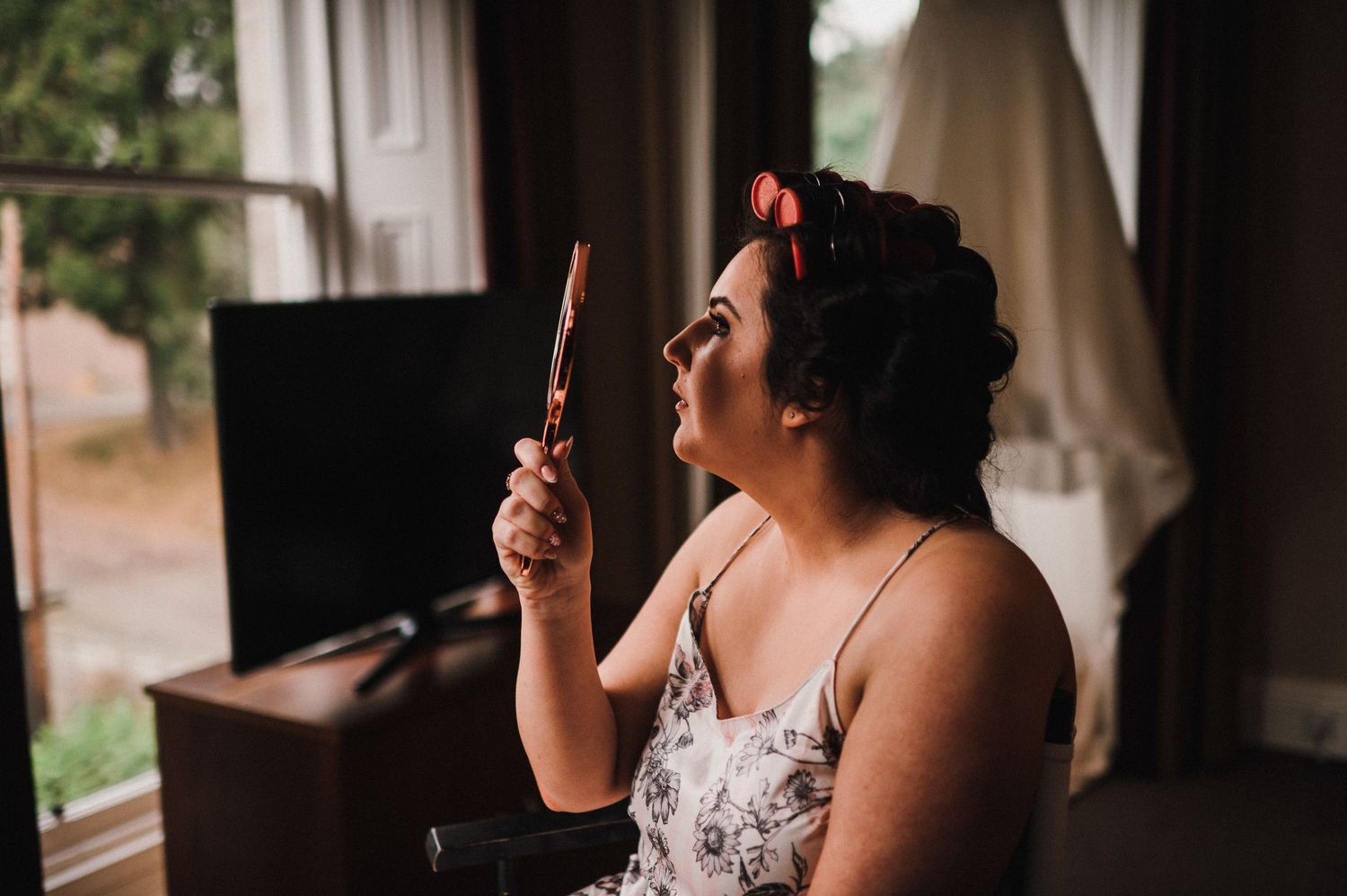 Person in floral dress and hair rollers looking at makeup mirror during wedding preparations.